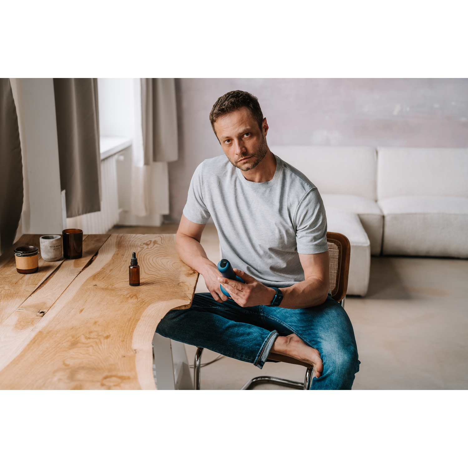 A man in a grey shirt and jeans sitting on a chair, holding a blue device.