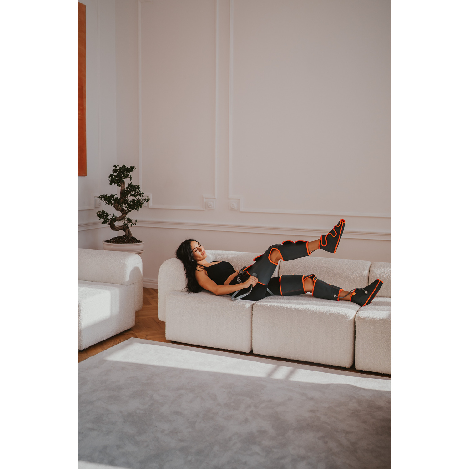 Woman on a couch wearing leg massagers, with plant and decor.
