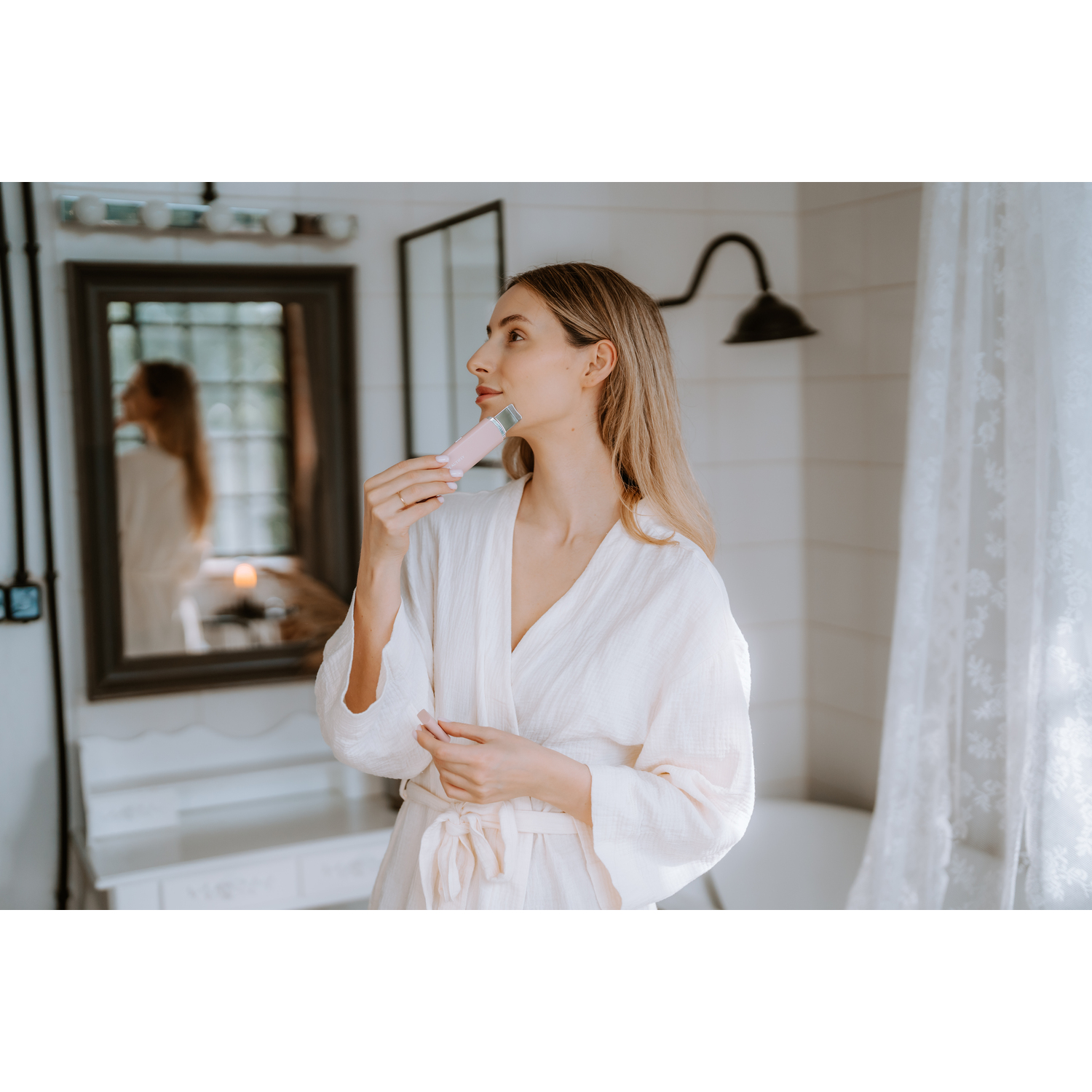 Woman in a white robe in a bathroom, using a pink skincare device, with mirror and sink visible.