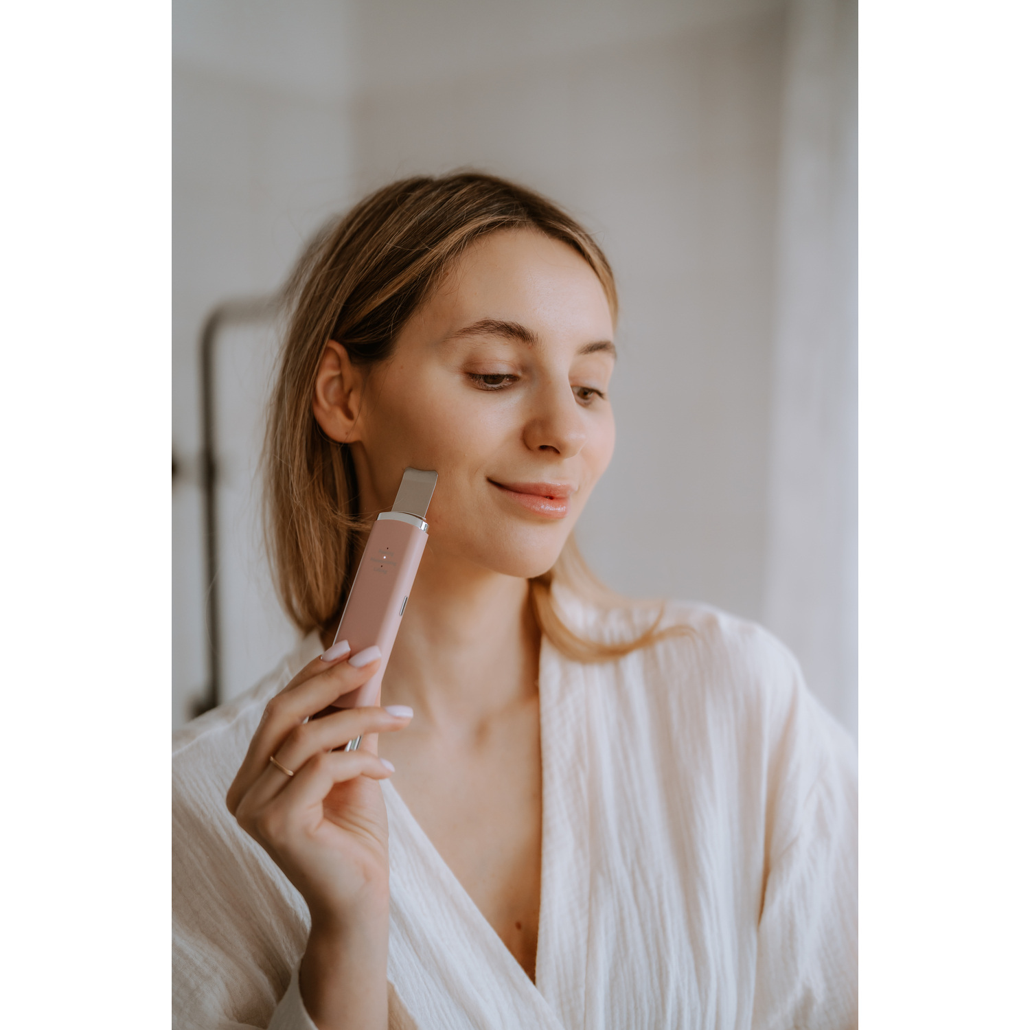 Woman in a robe applying a pink skincare device to her face, smiling, in a bathroom setting.
