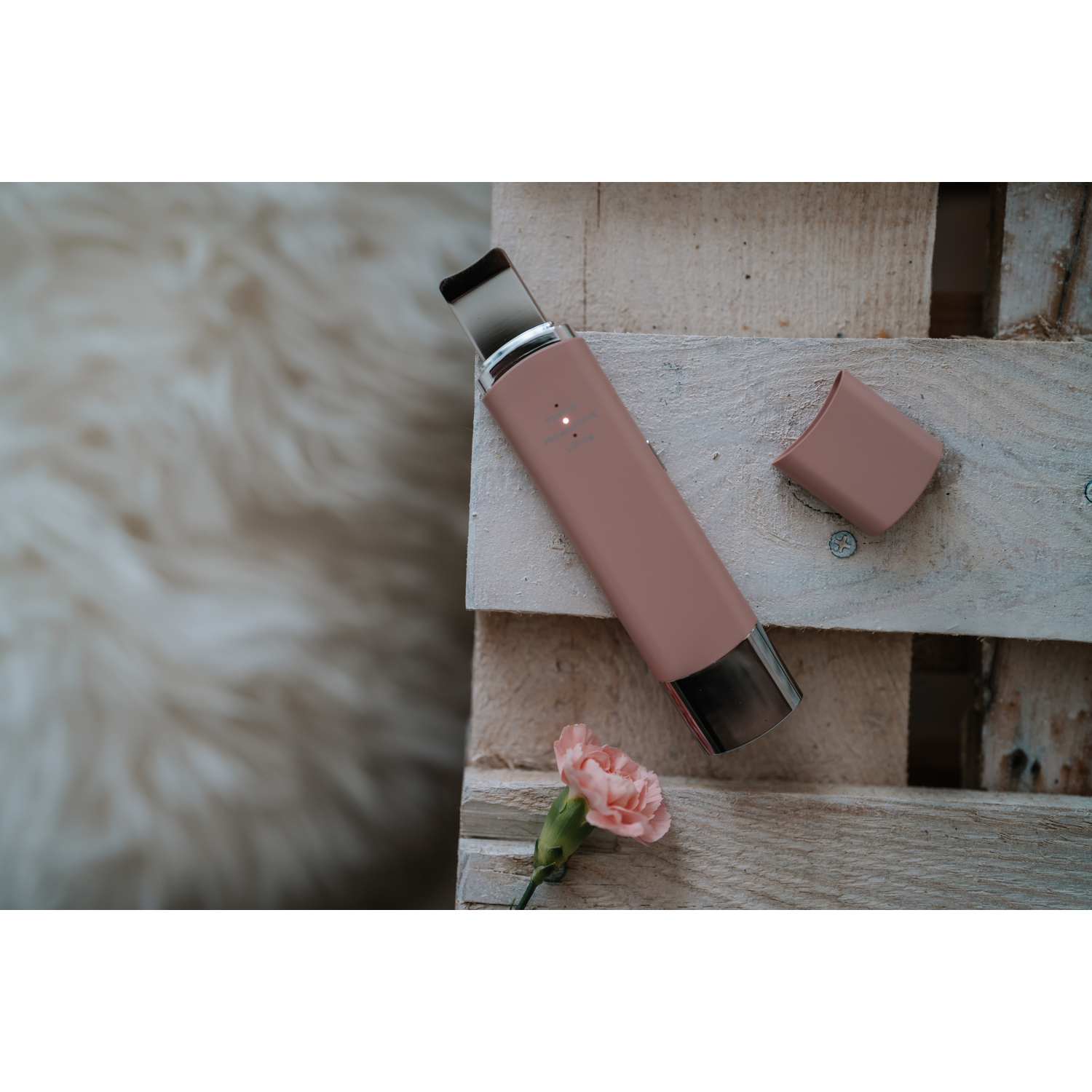 A pink facial cleansing device on wood, a lid, and a pink carnation flower.