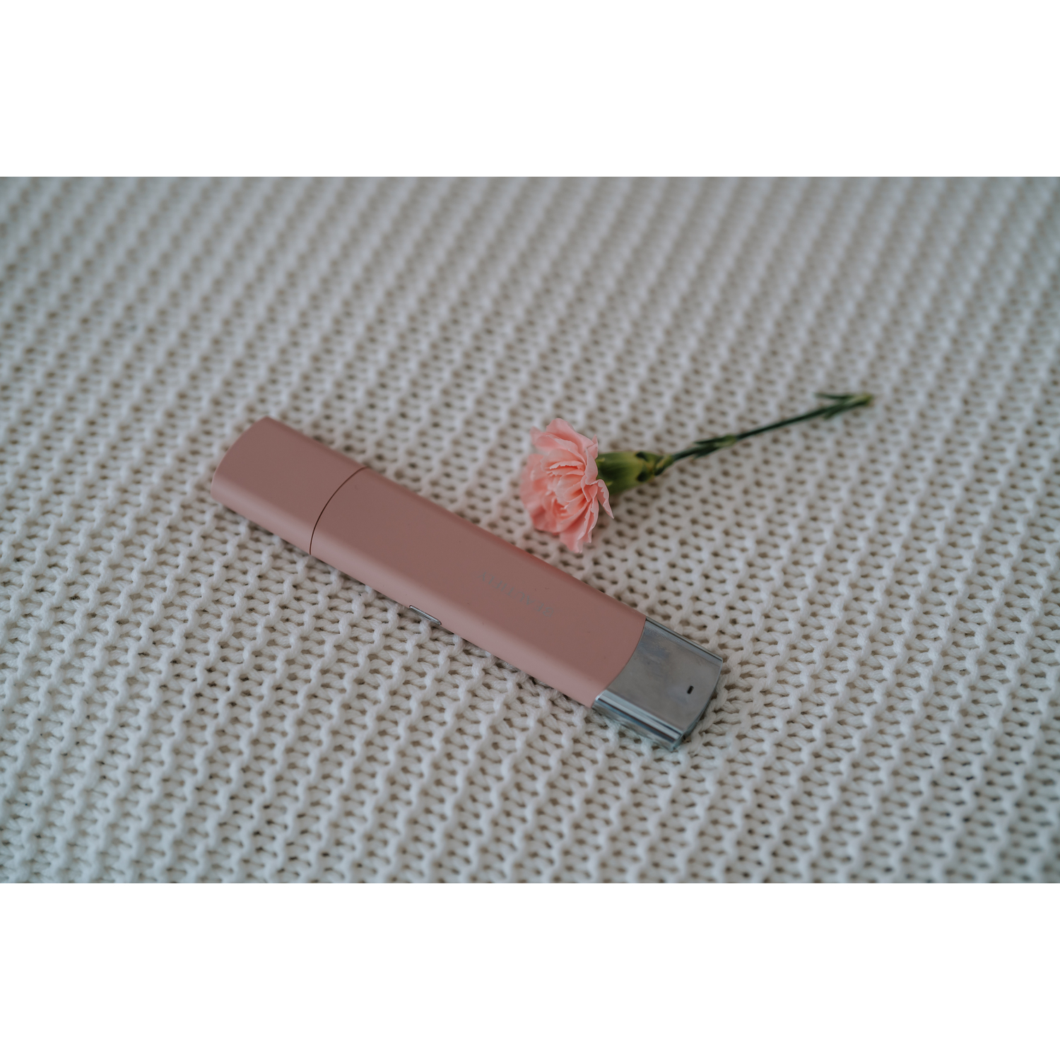A pink lipstick and a pink carnation flower on a white, textured, knitted blanket. The lipstick is closed.