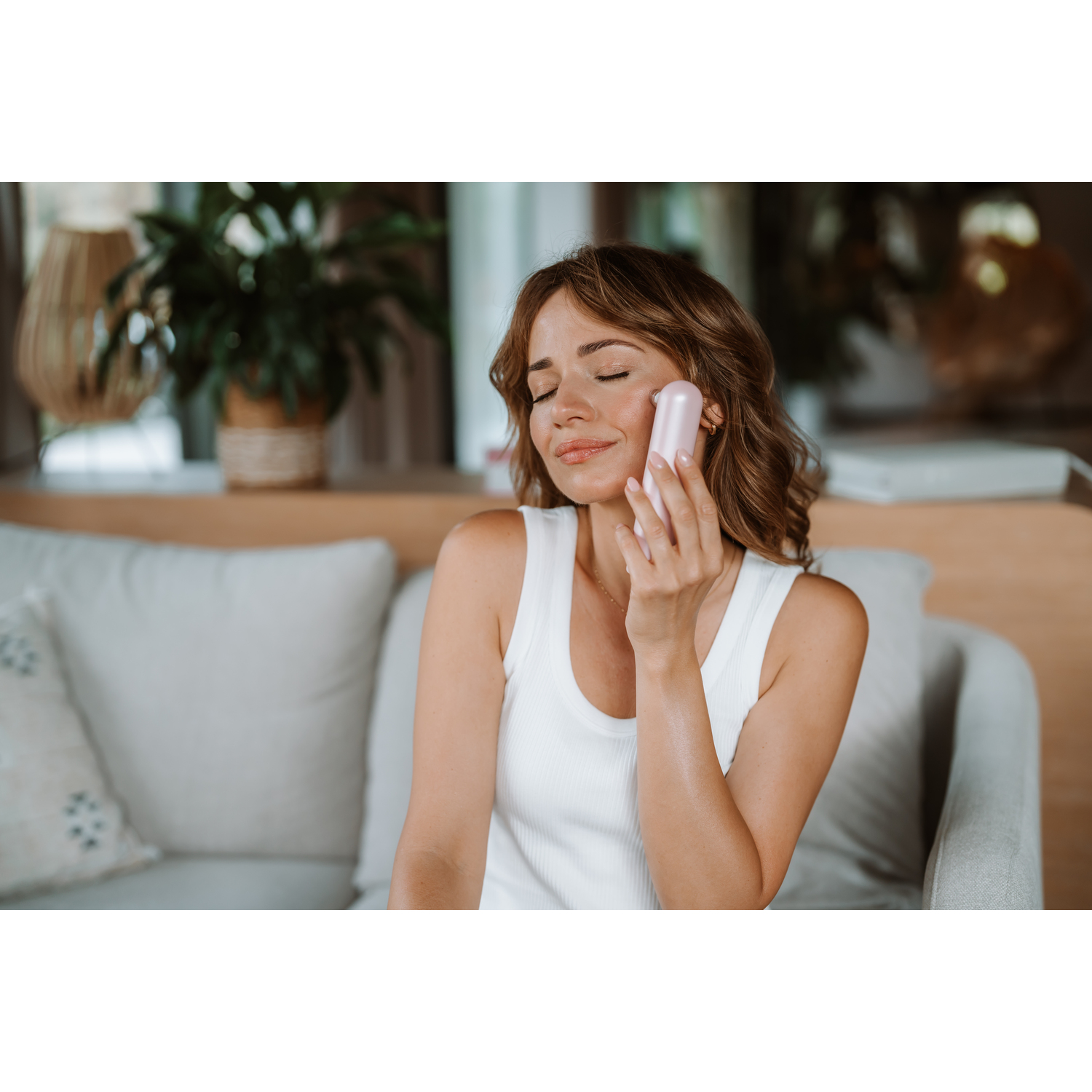 Woman using pink beauty device on face, eyes closed, in home setting. 