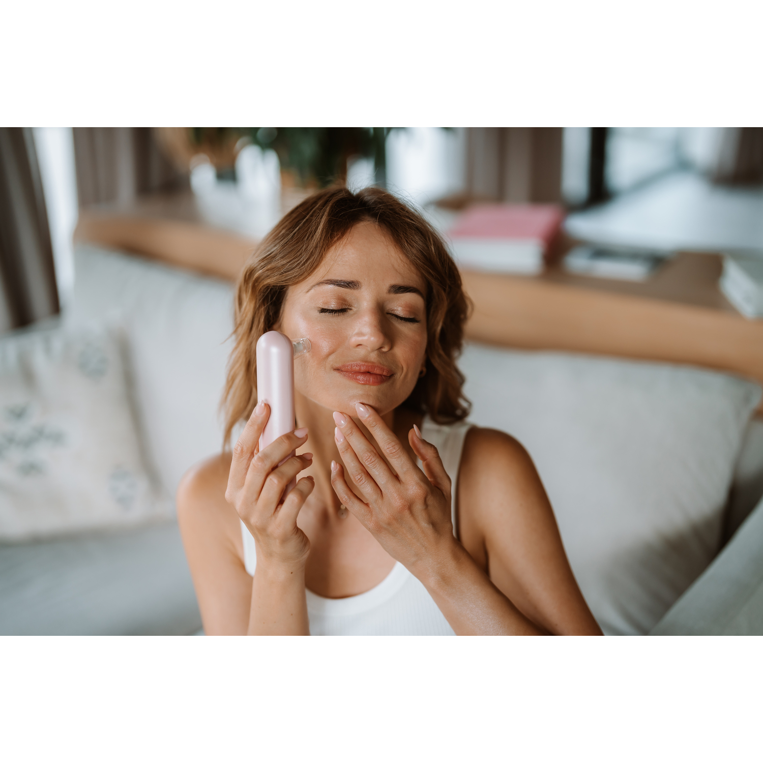 Woman holding a pink beauty tool, closed eyes, touching her face. Interior scene with pillows and window.
