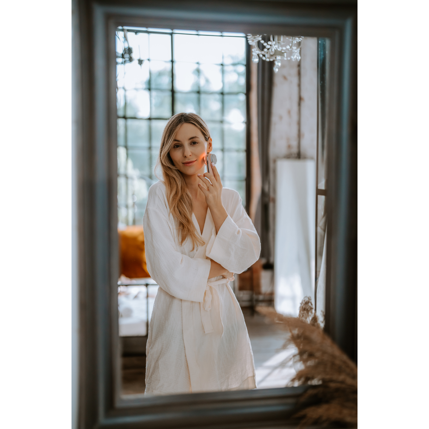 Woman in robe uses facial device, reflected in mirror. Background has a window and chandelier.