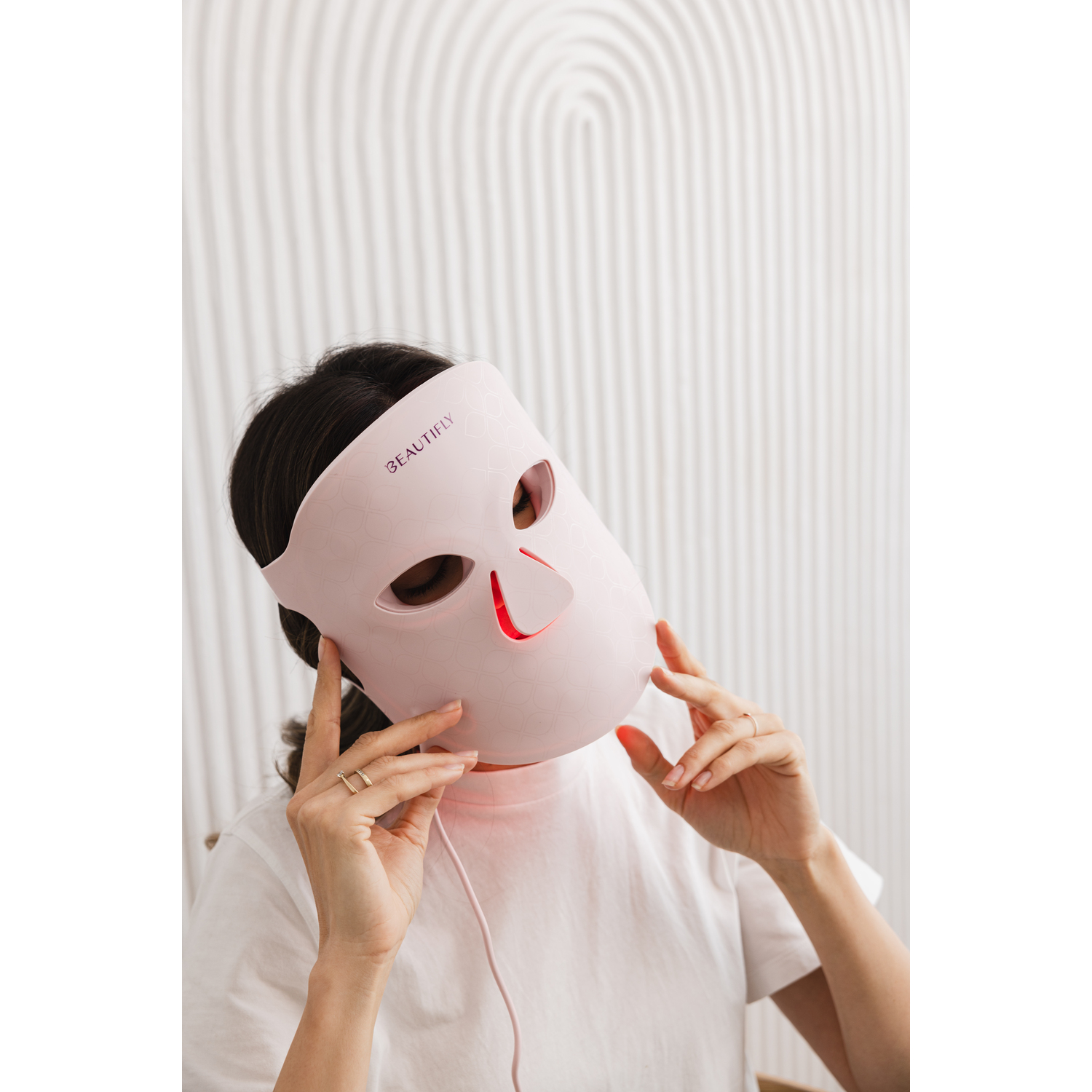 A woman wearing a pink LED face mask, hands touching the mask, against a white textured background.