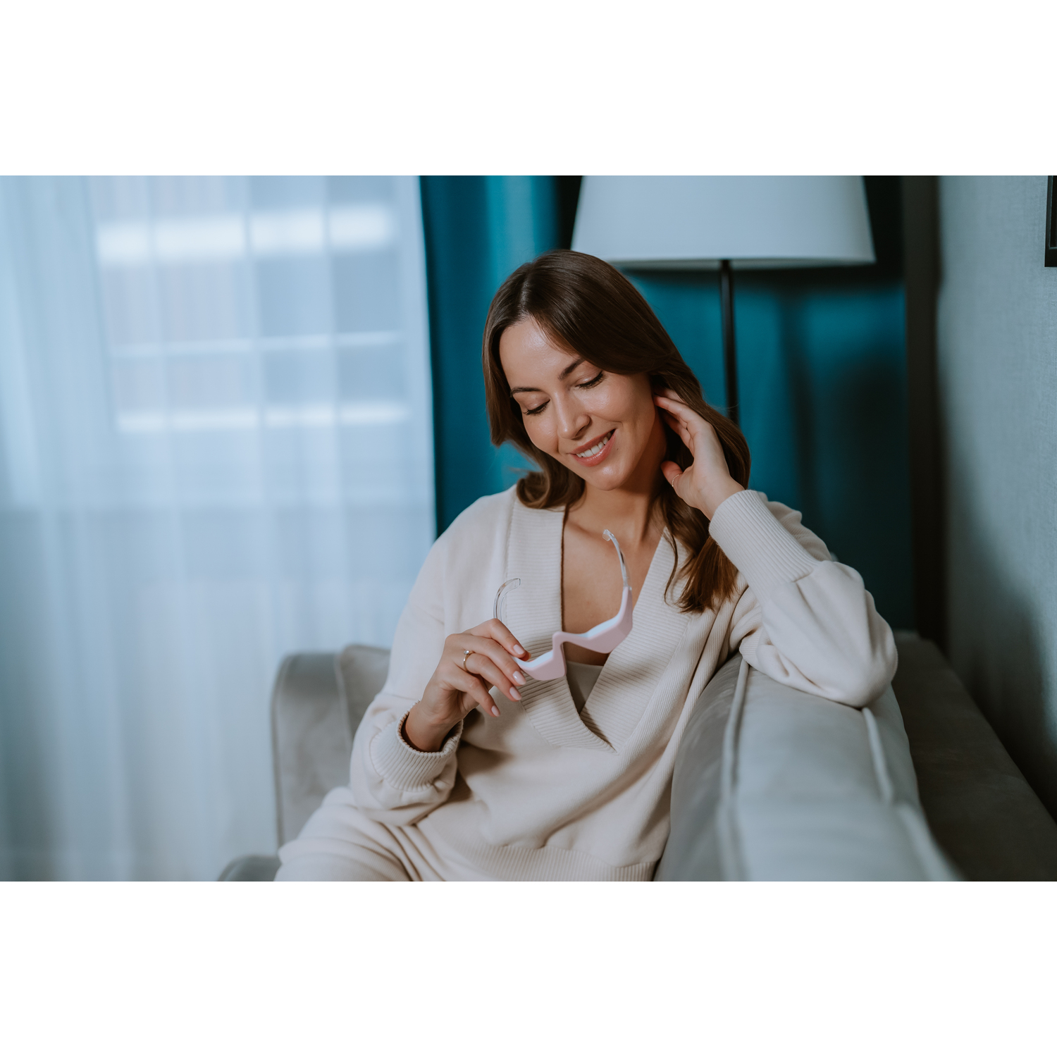 Woman holding pink eye massager, smiling, wearing white outfit on couch.