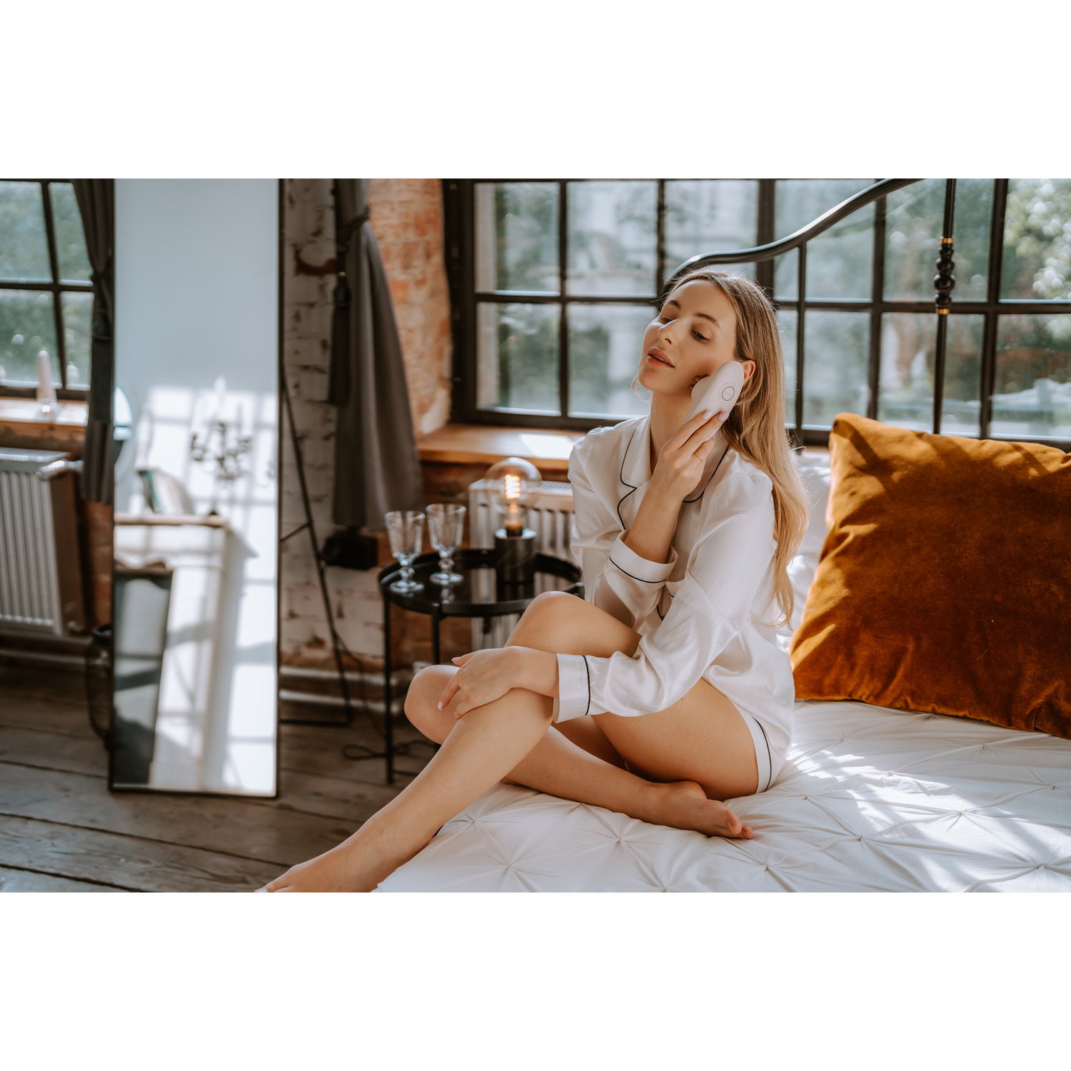 Een vrouw in een witte outfit gebruikt een gezichtsapparaat terwijl ze op een bed met kussens zit.