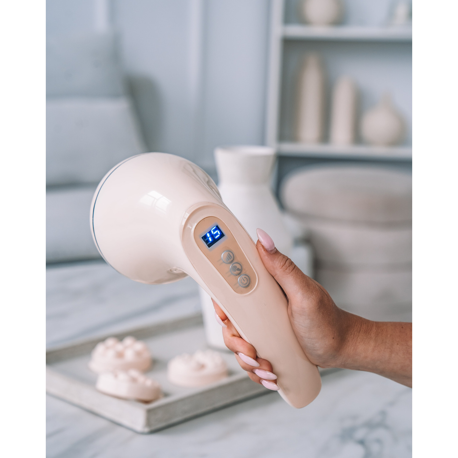 A woman holding a beige massage device with a display, on a tray with massage attachments.
