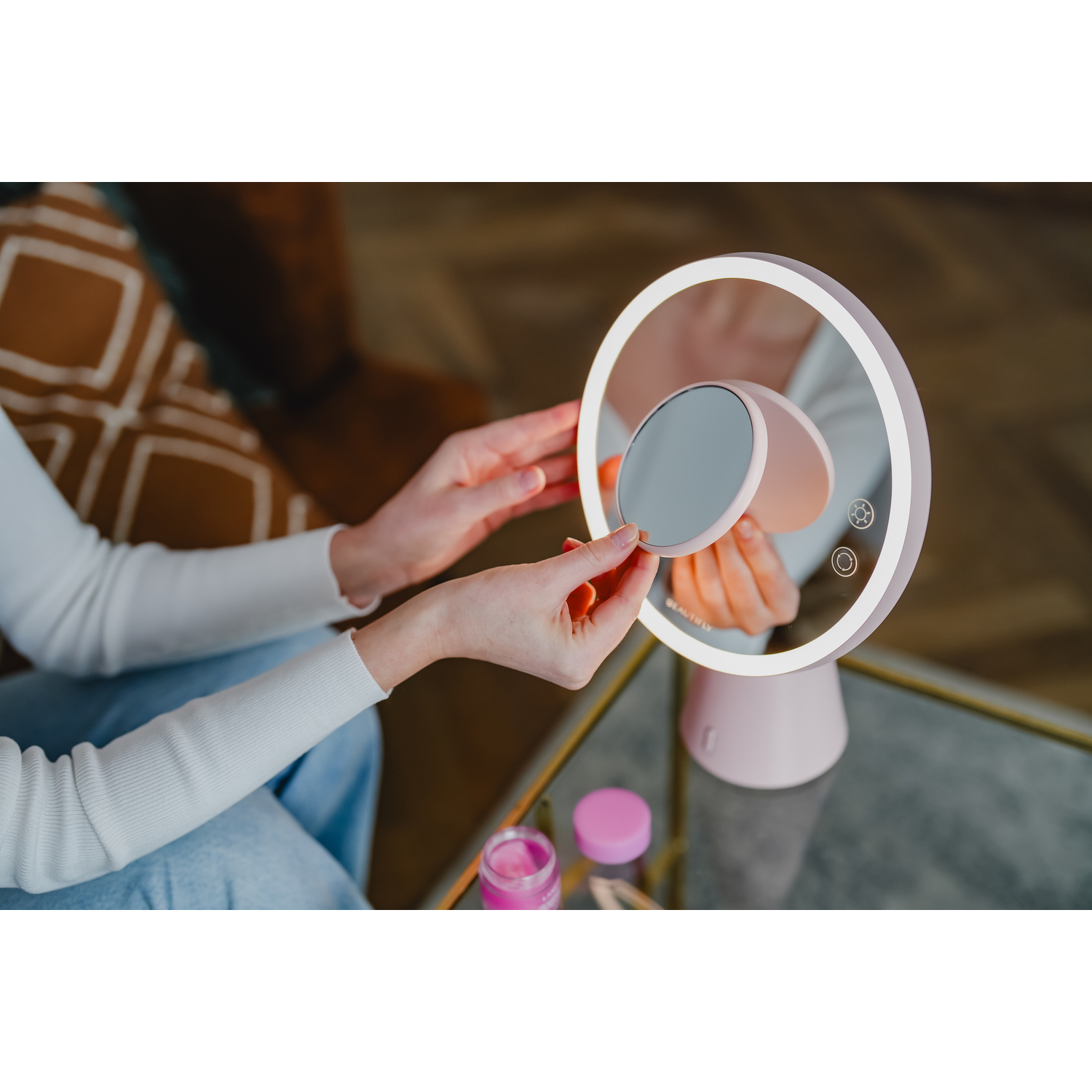 A woman's hands are adjusting the makeup mirror. Makeup items are on the table.