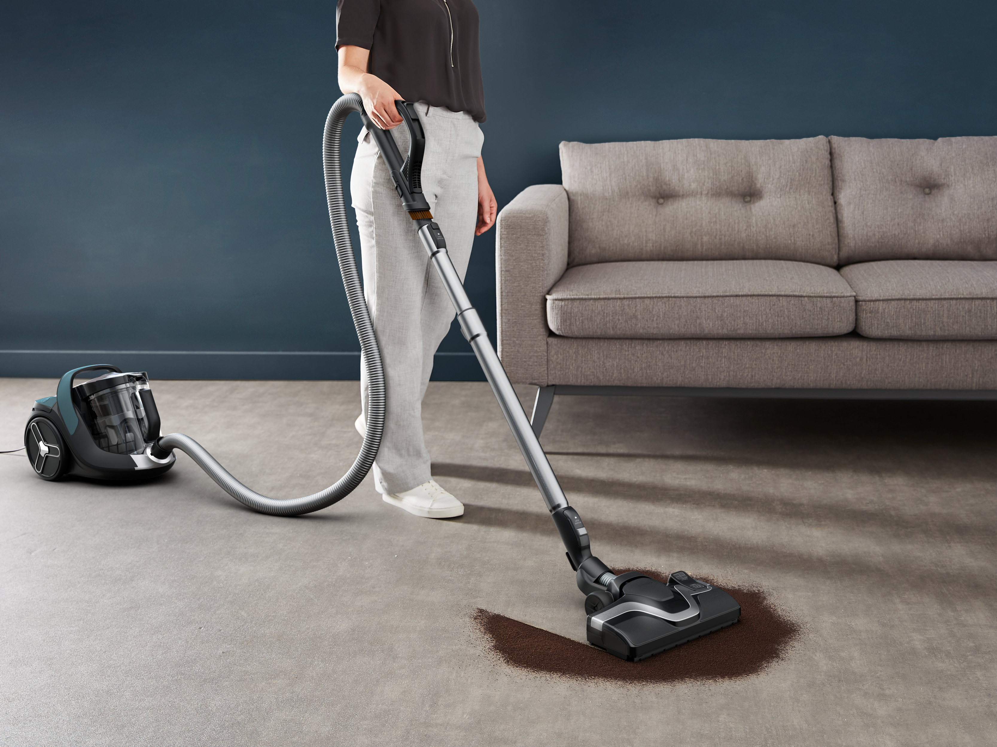Frau, die einen Teppich mit einem Staubsauger saugt, graues Sofa im Hintergrund.