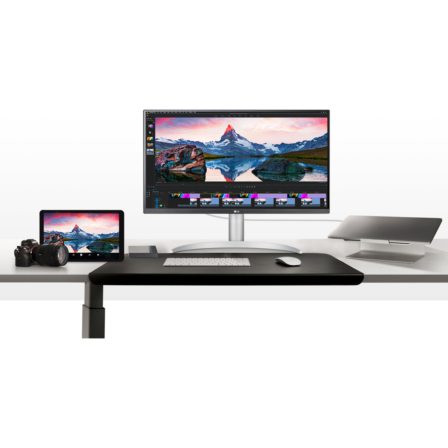 A desk setup features a monitor, tablet, laptop, keyboard, mouse, and camera. The monitor displays a mountain landscape.
