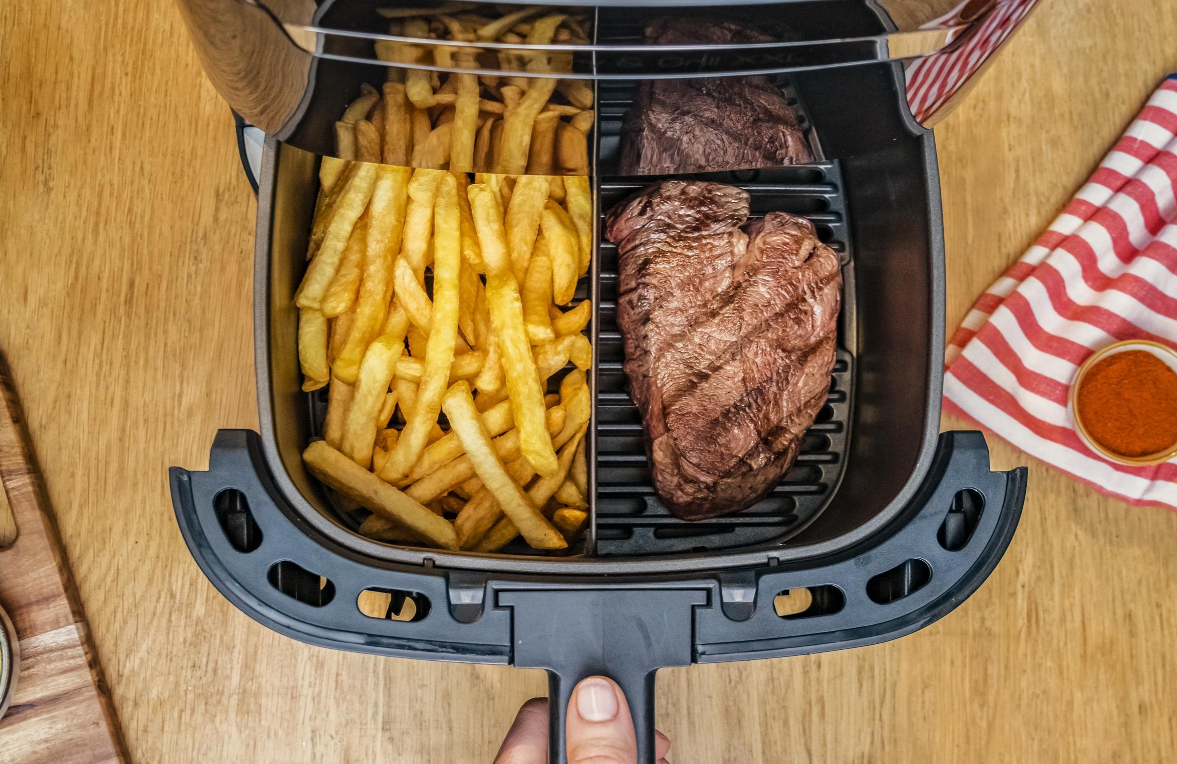 Friggitrice ad aria con patatine e bistecca. Patatine a sinistra, bistecca a destra. Sfondo tavolo in legno.