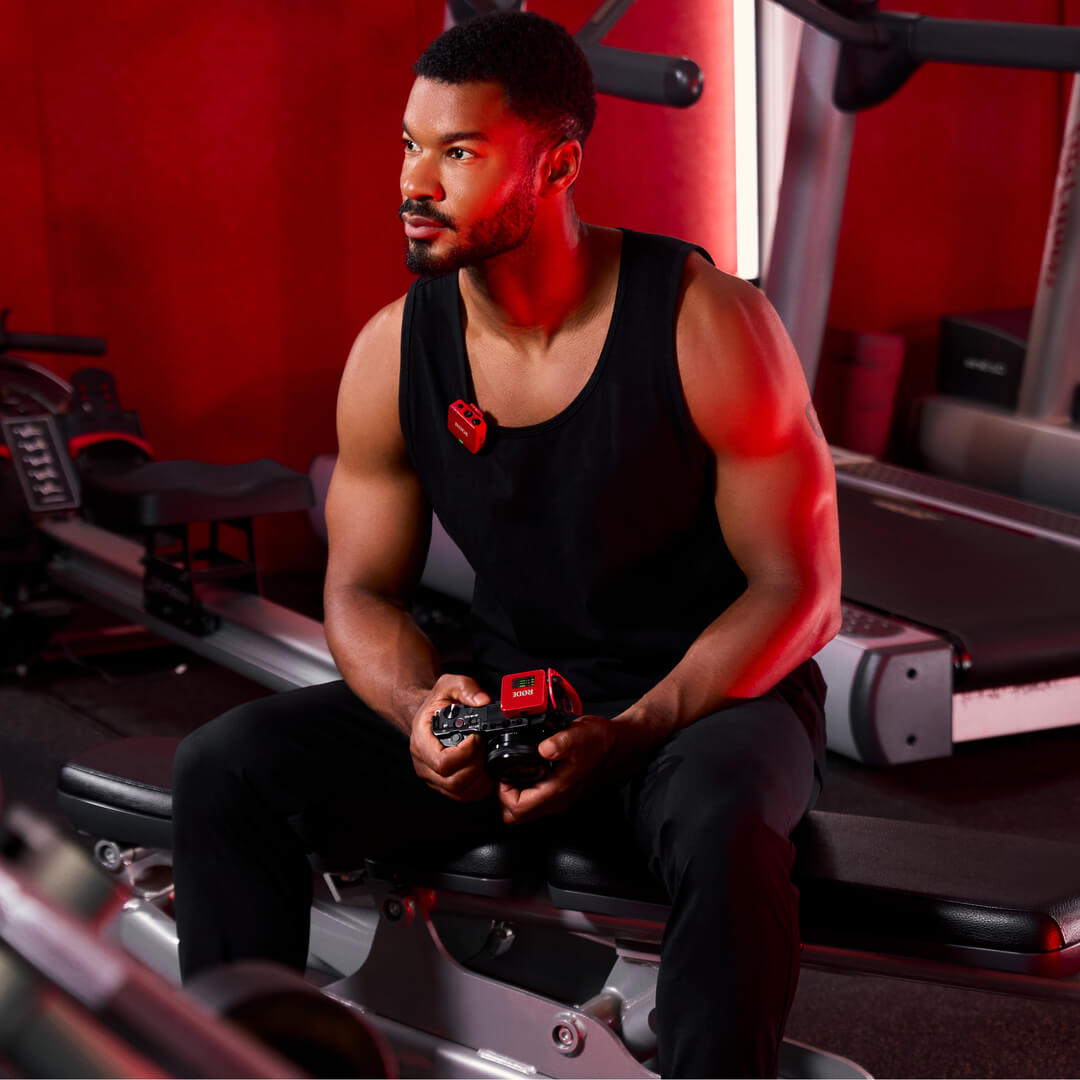 Homme en tenue de sport avec un microphone rouge, assis sur un banc avec des équipements de gym.