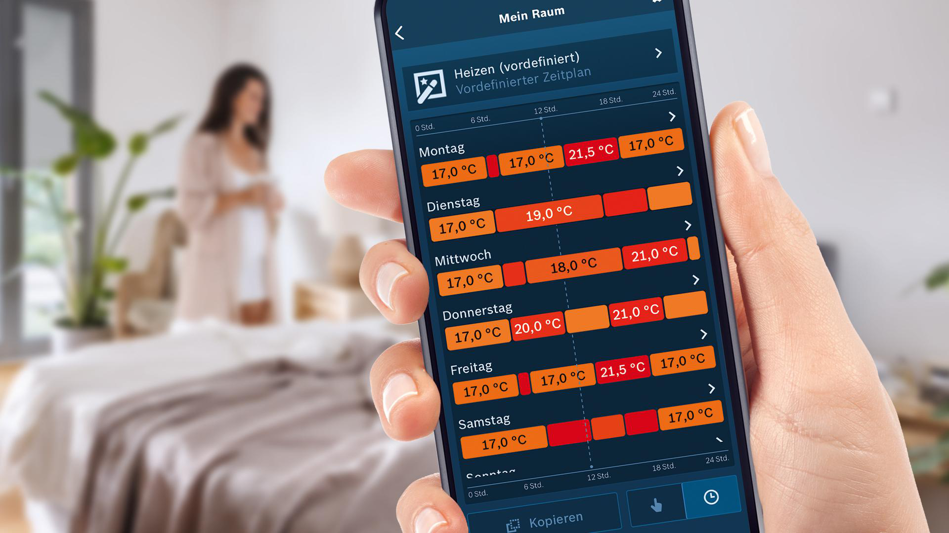 Hand hält Smartphone mit Heizplan. Frau im Bademantel im Hintergrund. Bett im Vordergrund.