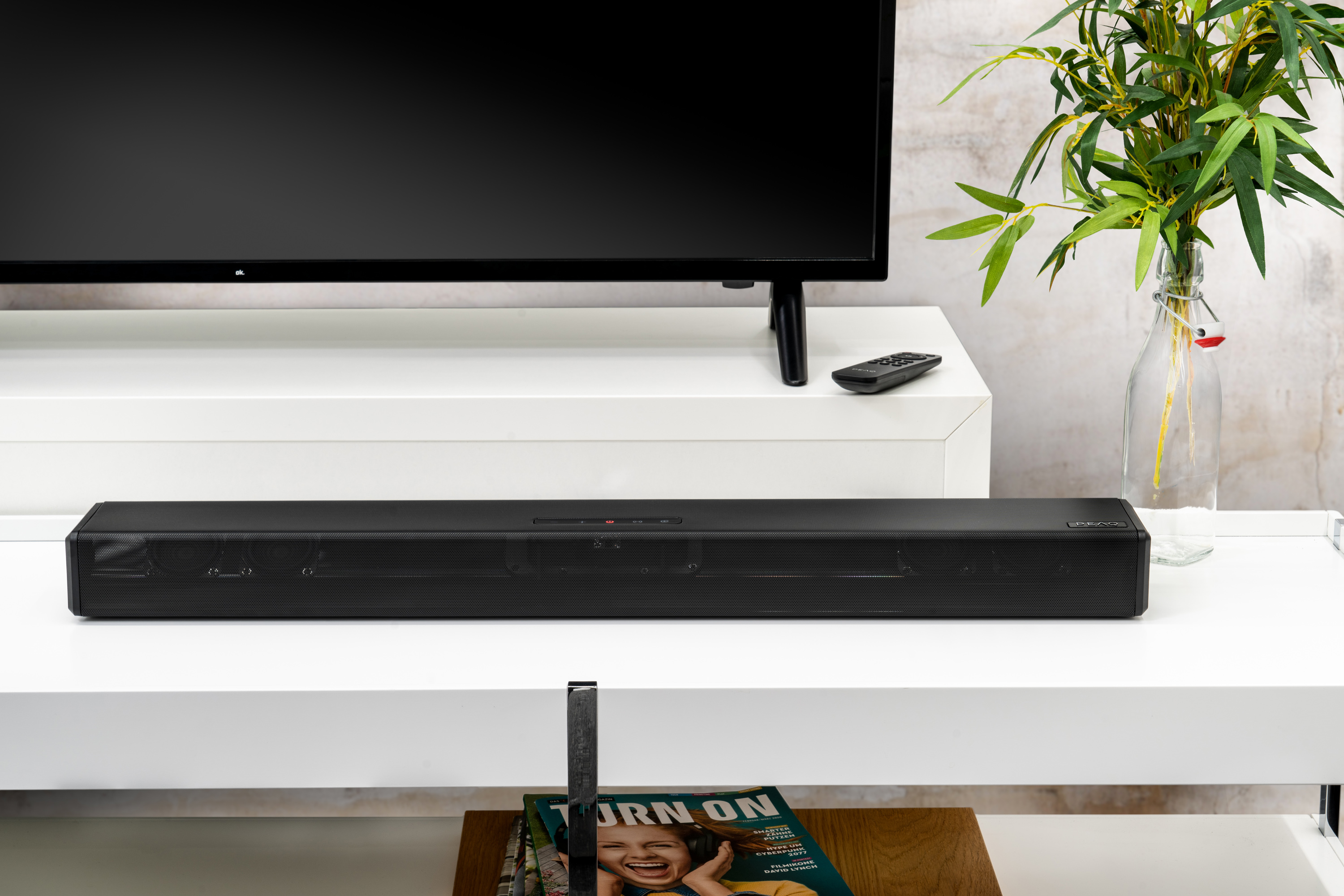 Une TV est sur une étagère blanche. Barre de son, télécommande et magazines en dessous.