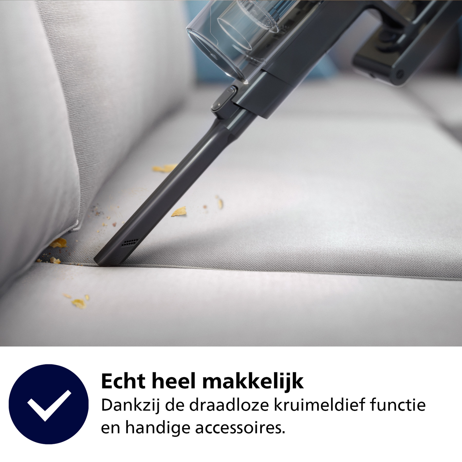 Een donkergrijze stofzuiger die een lichtgrijze bank schoonmaakt, met kruimels zichtbaar. De achtergrond is wazig.