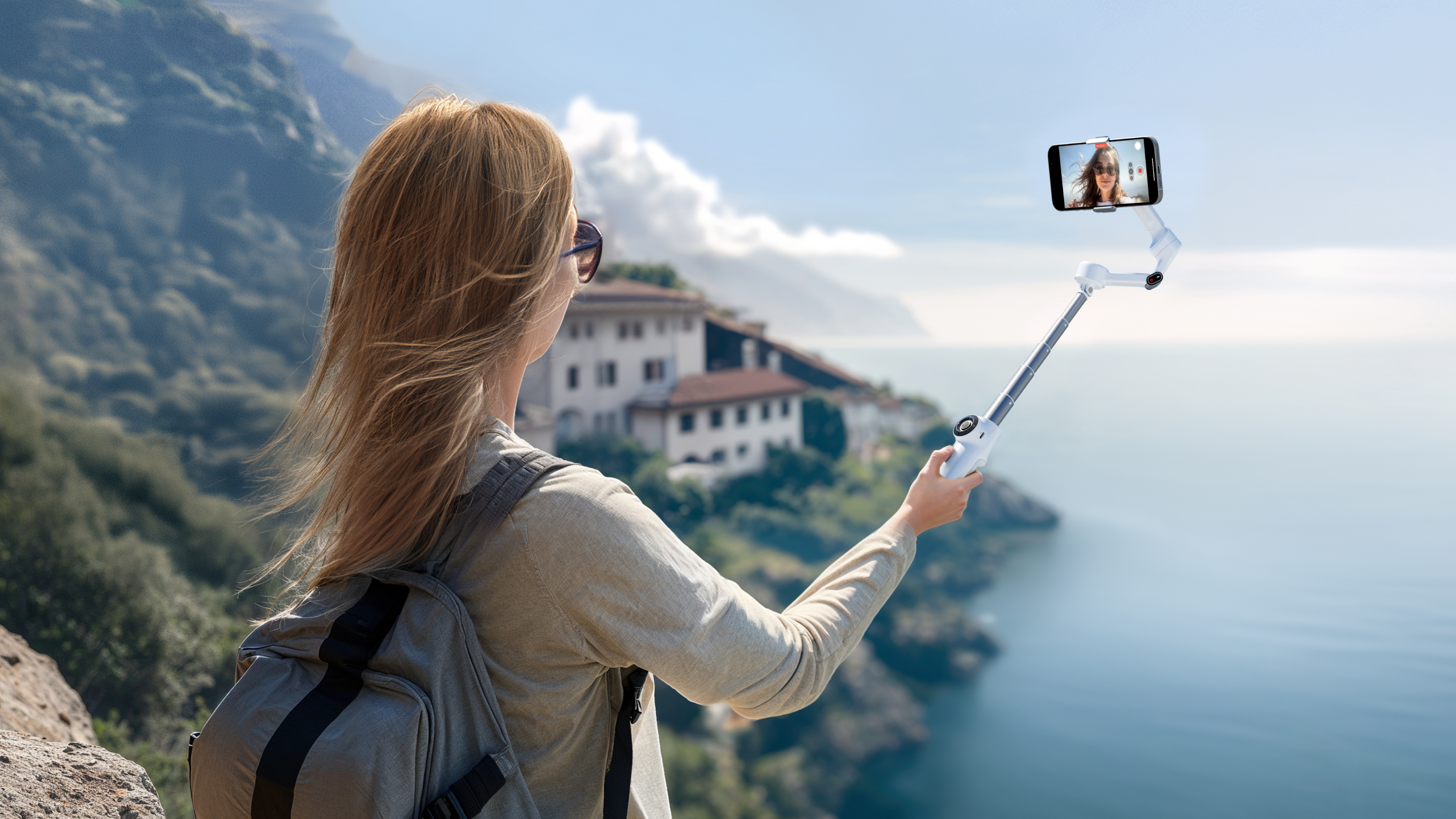 Femme avec perche à selfie, montagnes et mer en fond.