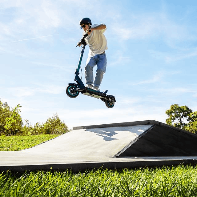 Persona en patinete saltando rampa en skatepark, cielo azul y hierba verde al fondo.