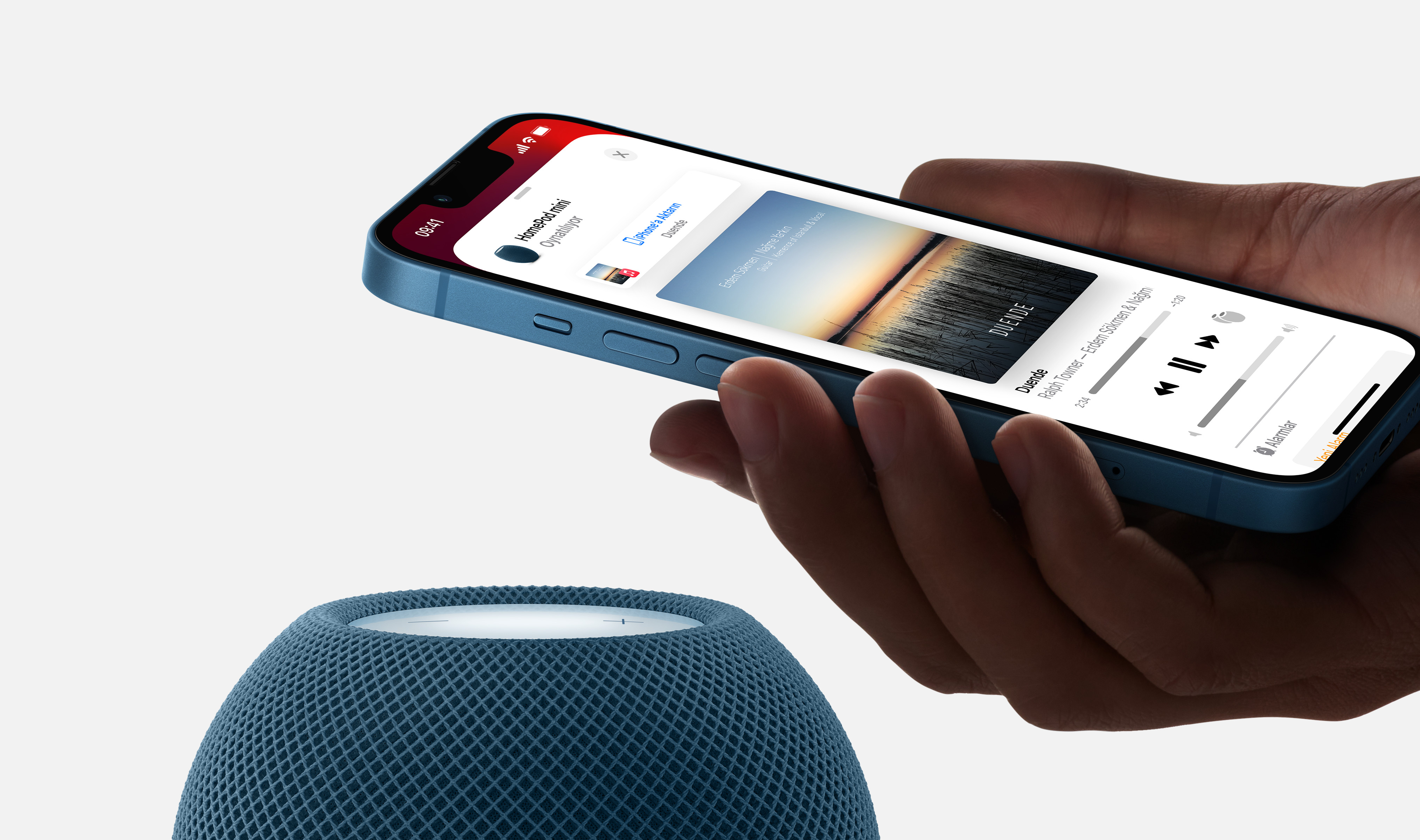 Mavi bir iPhone'u müzik kontrolleri gösterirken tutan bir el, önünde mavi bir hoparlör.