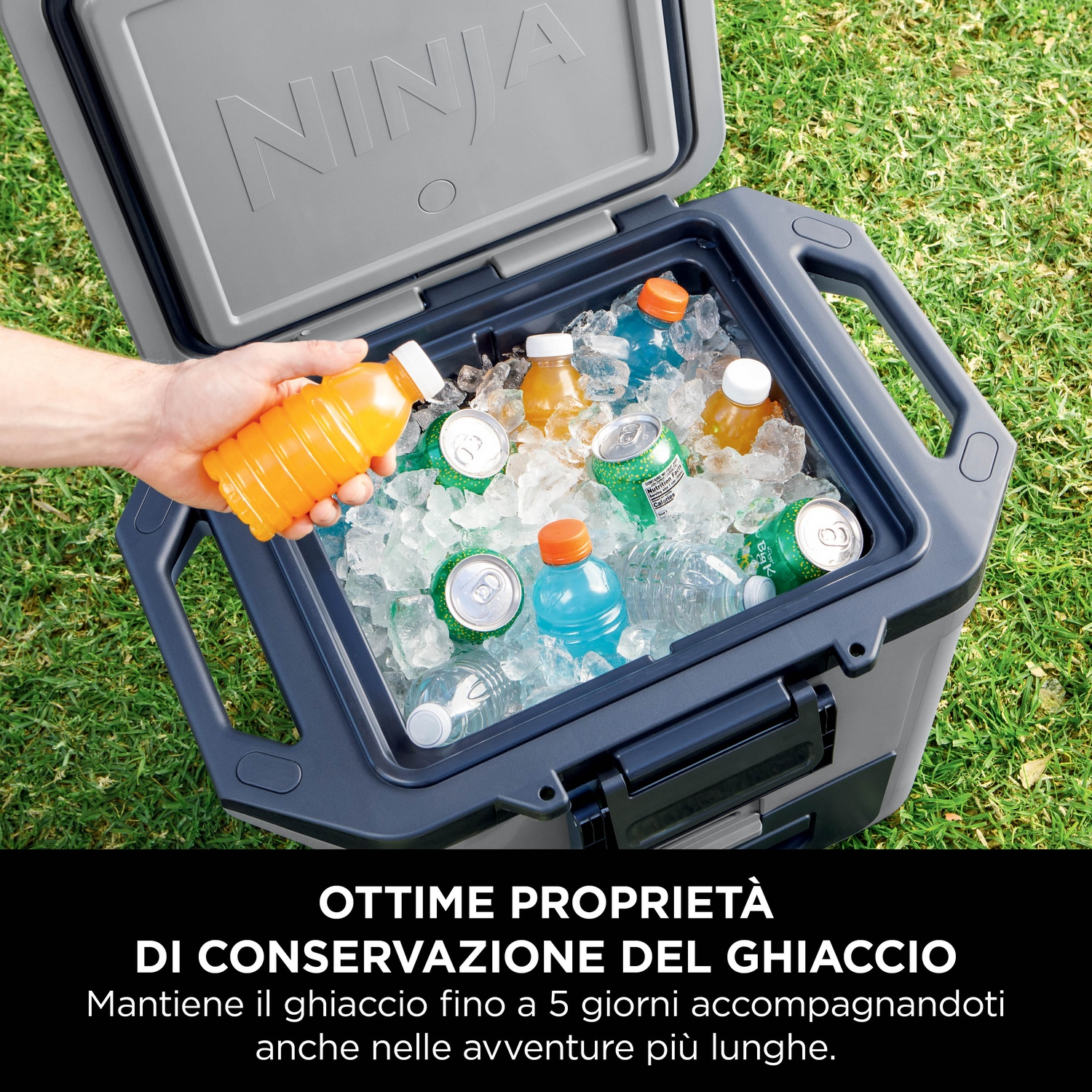Frigorifero aperto con bevande sul ghiaccio. Bottiglia arancione in mano. Erba verde sullo sfondo.