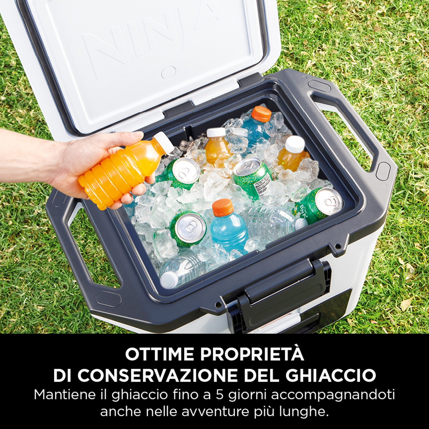 Un refrigeratore aperto sull'erba con ghiaccio, bevande e una mano che raggiunge una bevanda arancione.