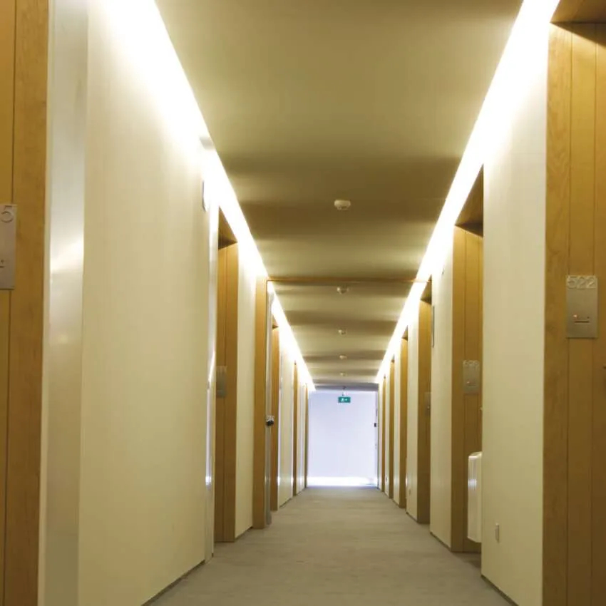 Jasny korytarz hotelowy z listwami świetlnymi na suficie i drewnianymi drzwiami po obu stronach.