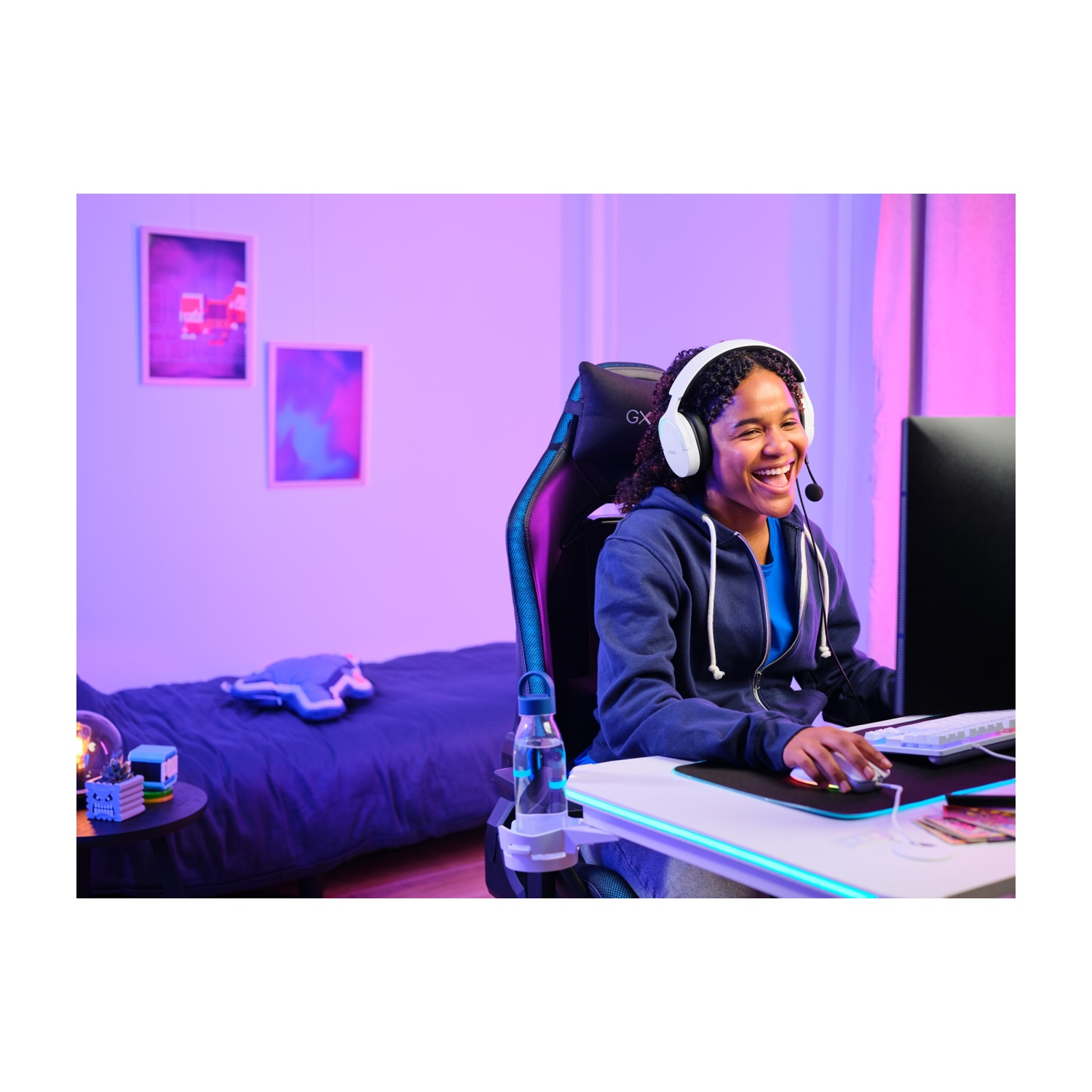 Una donna sorridente gioca al computer, con le cuffie. Sedia, scrivania e decorazioni da gioco sullo sfondo.