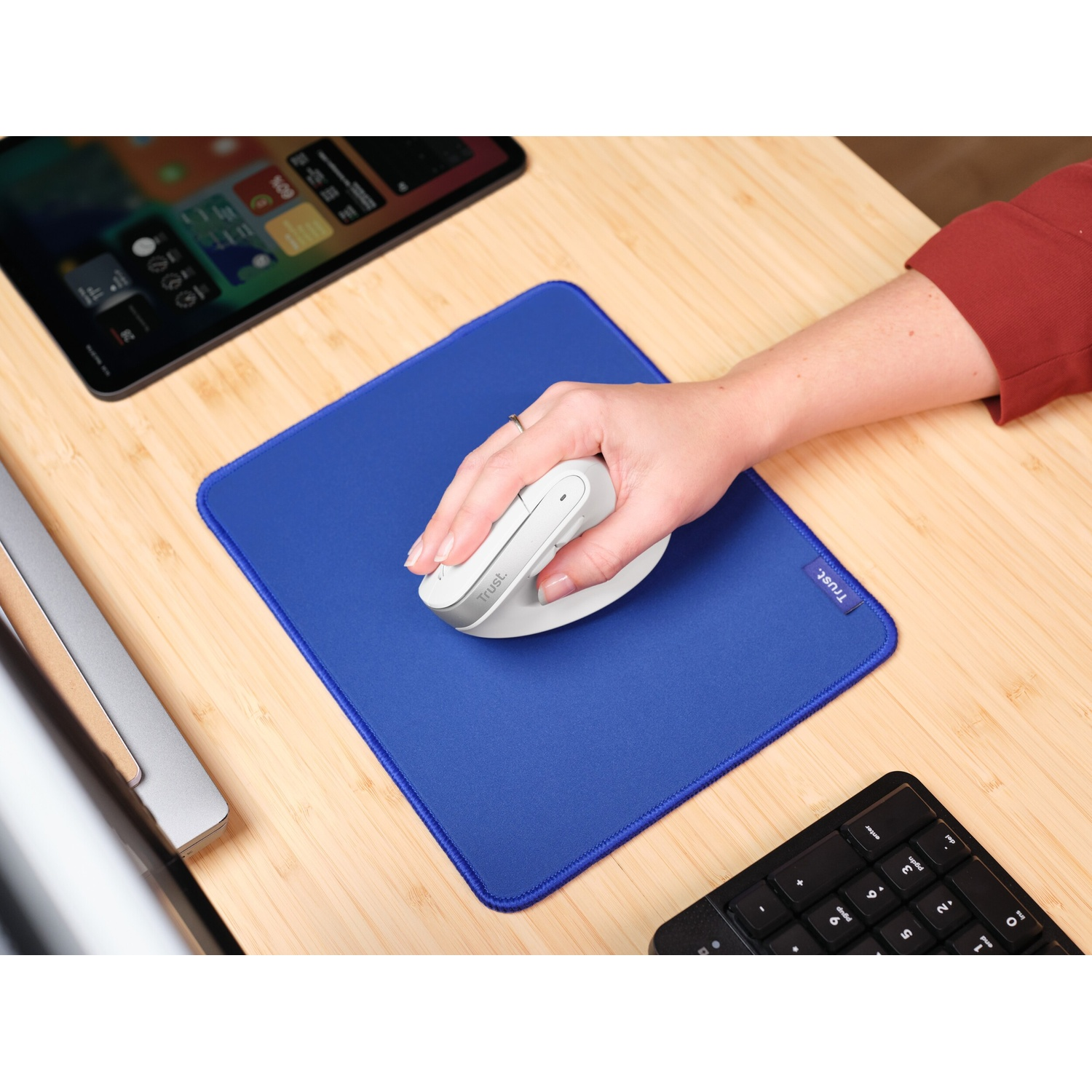 Una mano usa un mouse bianco su un tappetino blu. Un tablet e una tastiera sono sulla scrivania di legno.