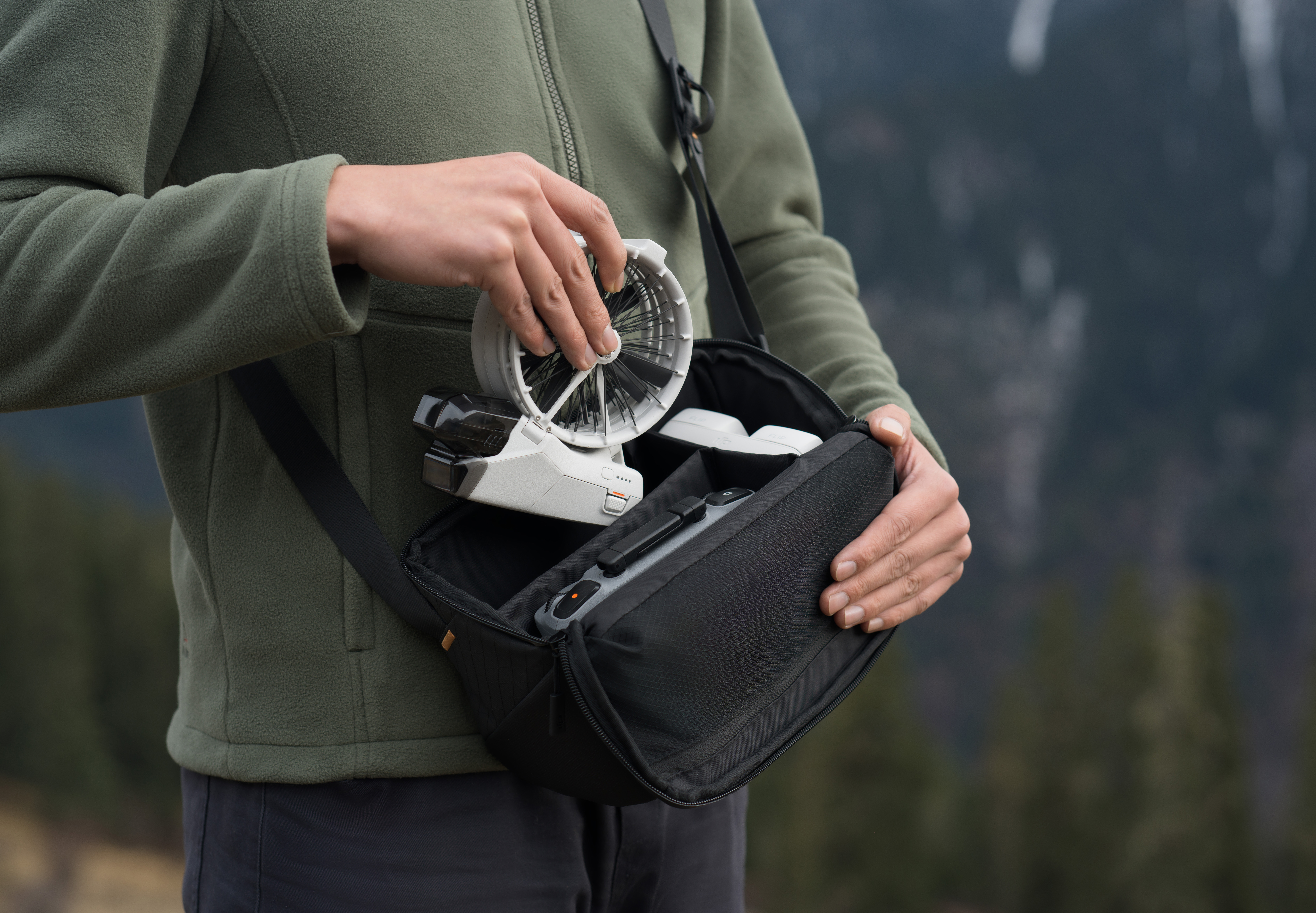 Eine Person öffnet eine schwarze Tasche mit einem kleinen Ventilator und anderen Gegenständen. Berge im Hintergrund.