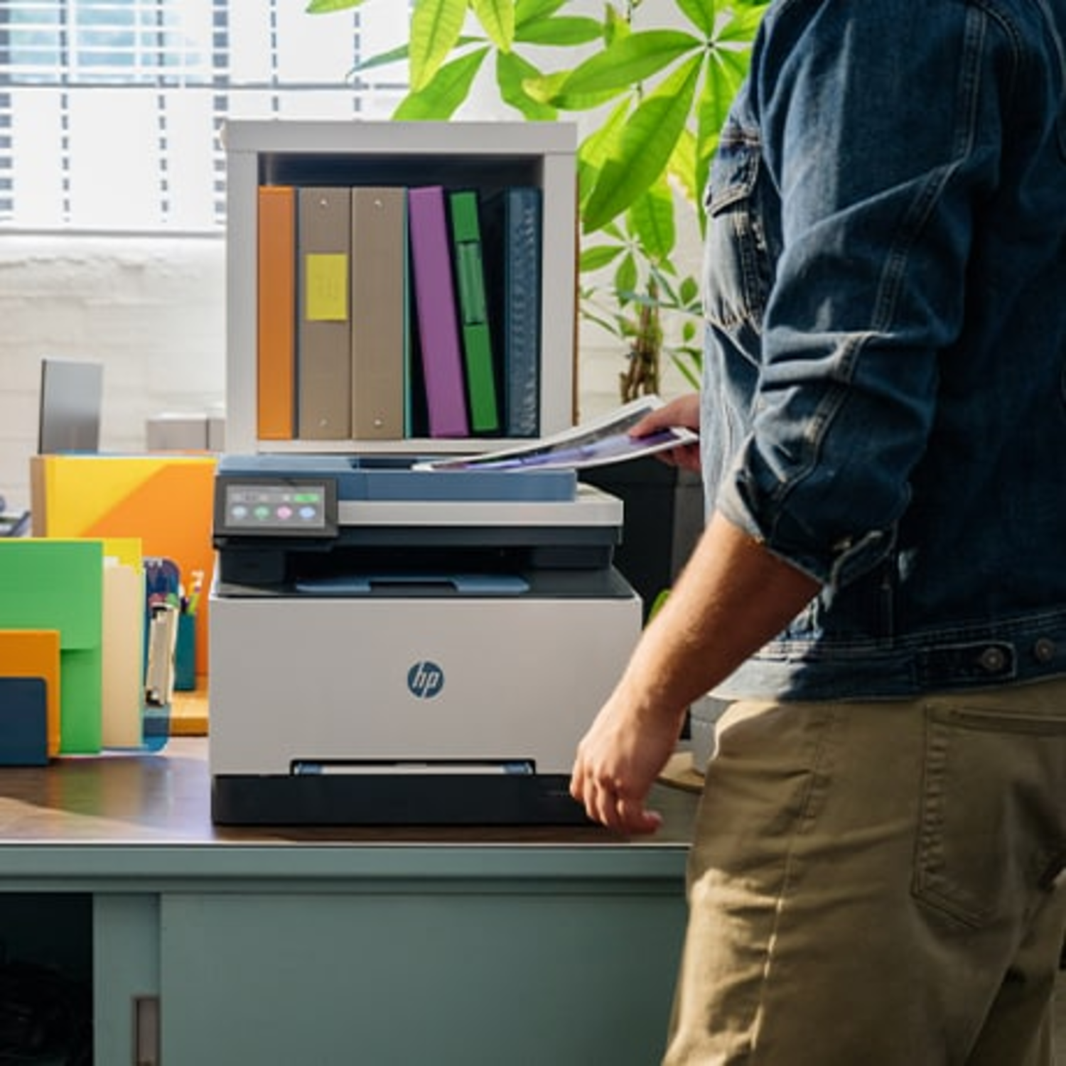 Een man in een spijkerjasje staat naast een HP-printer op een bureau.
