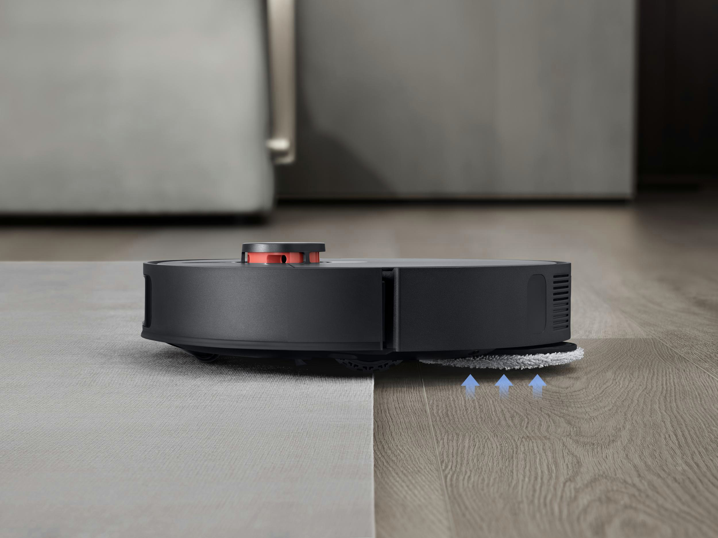 Ein schwarzer Roboter-Staubsauger reinigt einen Boden. Zeigt einen grauen Teppich und Holzboden. Pfeile zeigen die Reinigungsrichtung an.