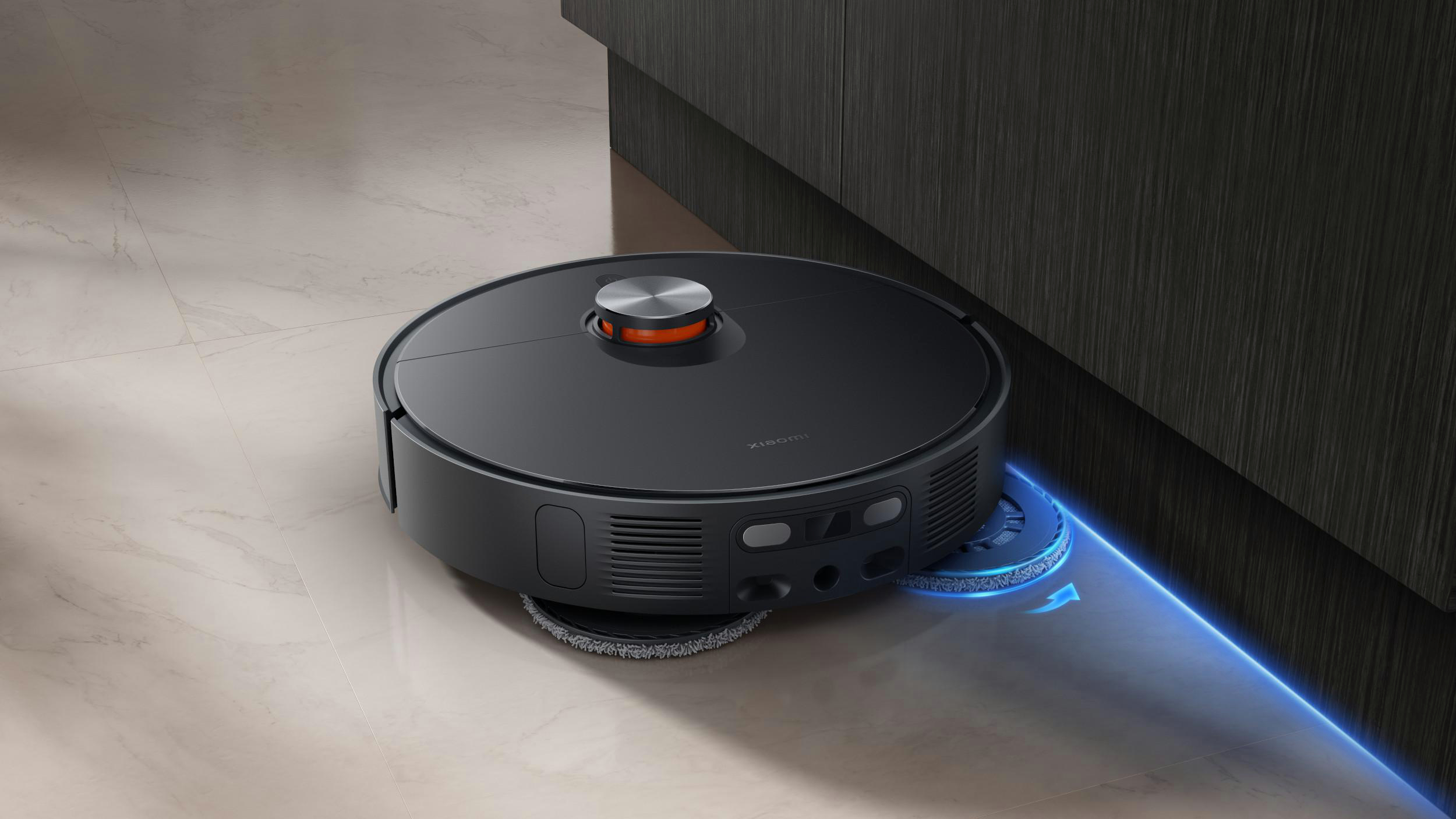 Ein schwarzer Roboter-Staubsauger mit orangefarbenen Akzenten, der einen weißen Fliesenboden in der Nähe eines dunklen Schranks reinigt.