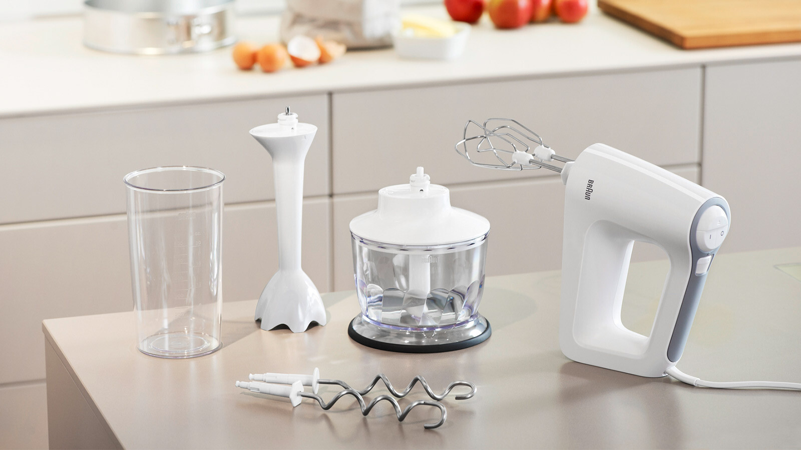 Weißer Handmixer mit mehreren Aufsätzen auf einer Theke. Zutaten sind im Hintergrund.