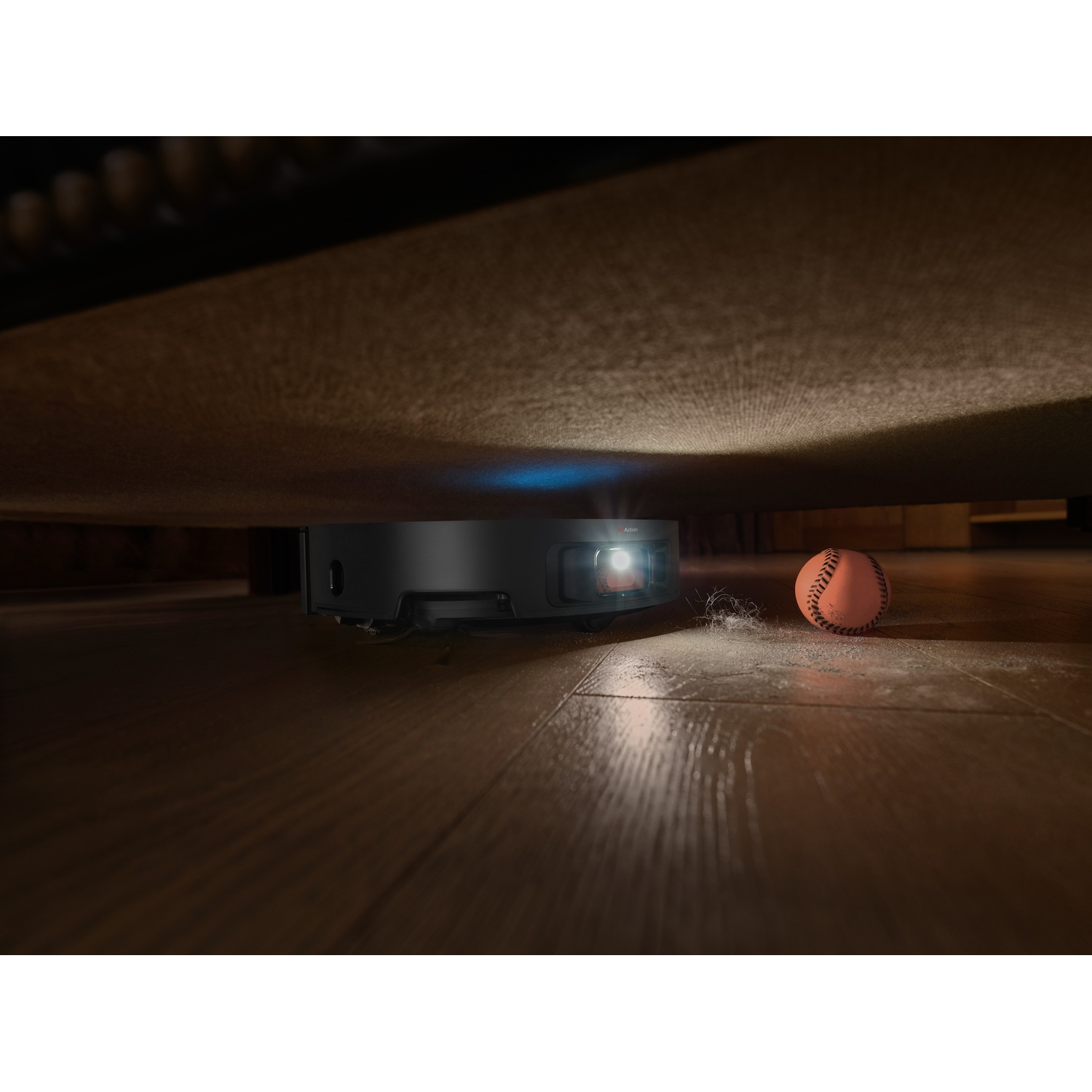 Aspirapolvere robot che pulisce sotto un divano. Una palla giocattolo è sul pavimento di legno.