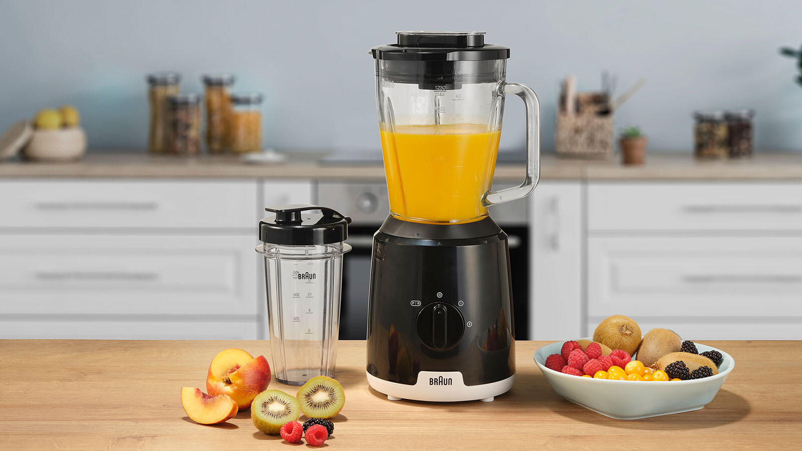 Mixer mit orangener Flüssigkeit. Früchte wie Kiwi, Pfirsich und Himbeeren auf dem Tisch.