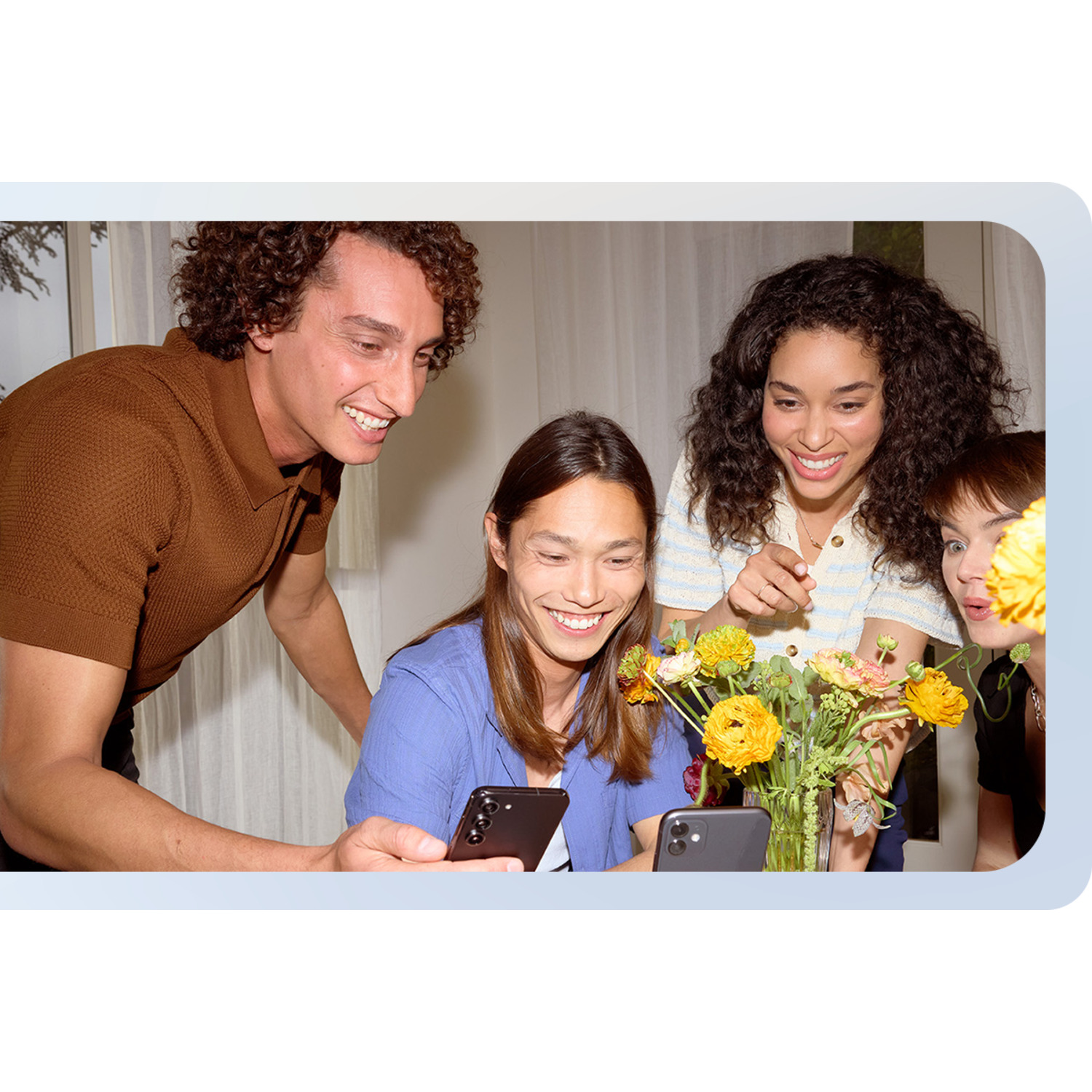 Vier mensen kijken naar twee telefoons, glimlachen, met een bloemenvaas.