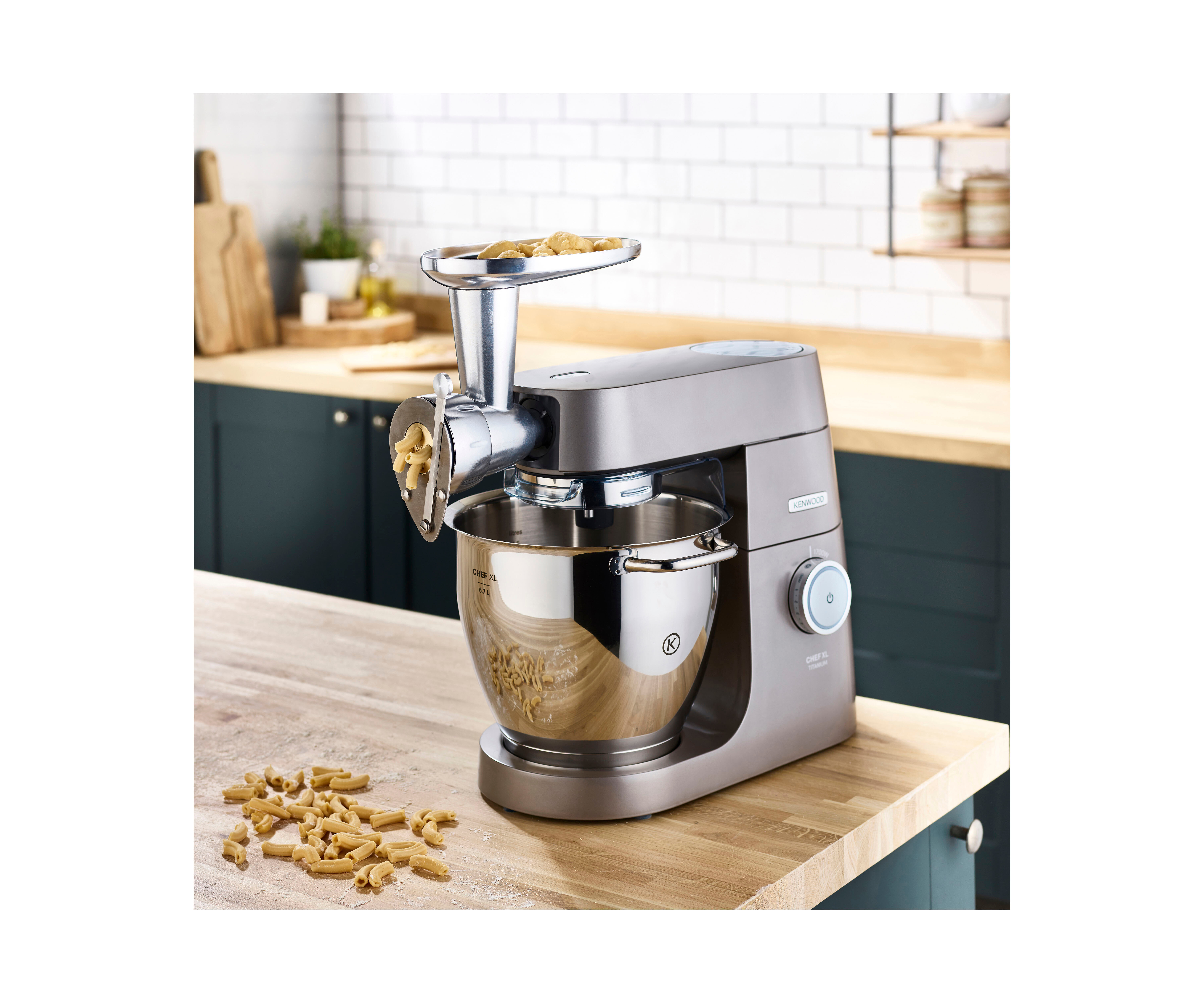 Grauer Kenwood Chef XL mit Nudelmaschine. Nudeln kommen heraus. Holztisch und grüne Schränke im Hintergrund.