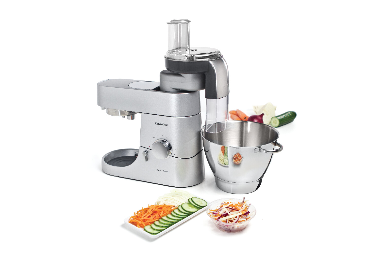 Silberfarbene Küchenmaschine mit Schüssel und Gemüse auf weißer Fläche.