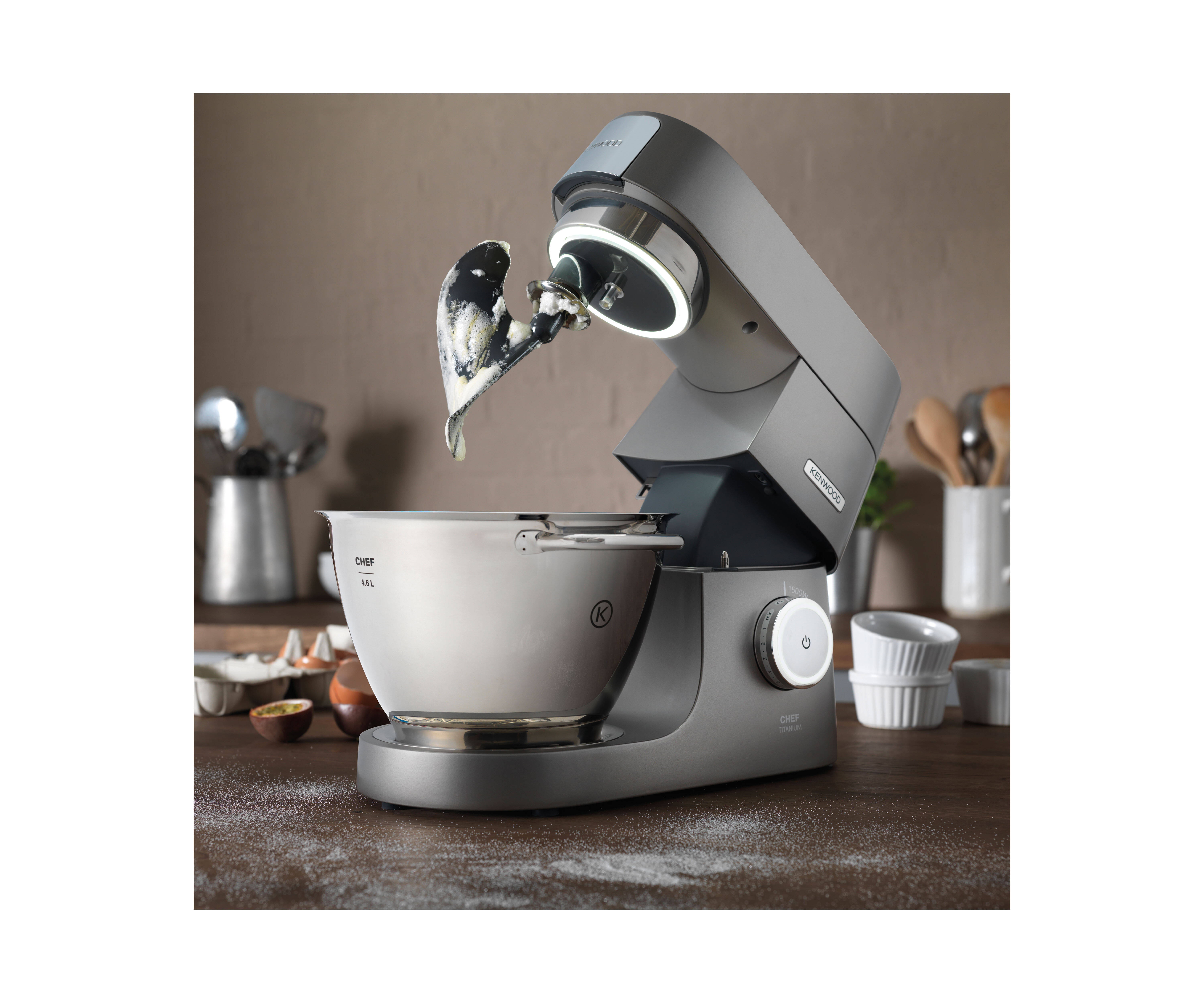 Ein silberner Küchenmixer mit Schüssel. Der Mixer steht auf einer Holzoberfläche. Mehl liegt auf der Oberfläche.