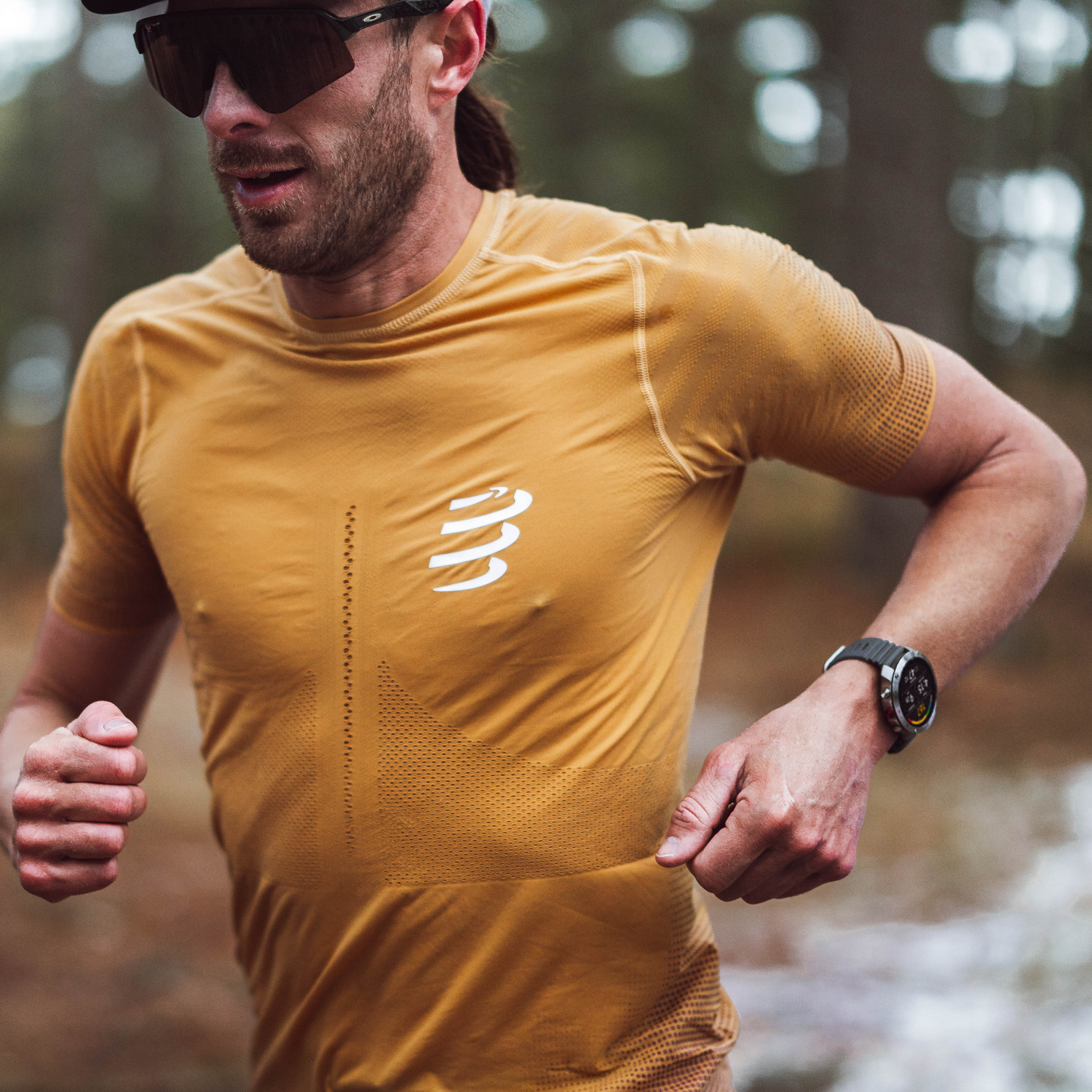 Hombre corriendo al aire libre, con gafas de sol y camisa amarilla. Fondo árboles borrosos.