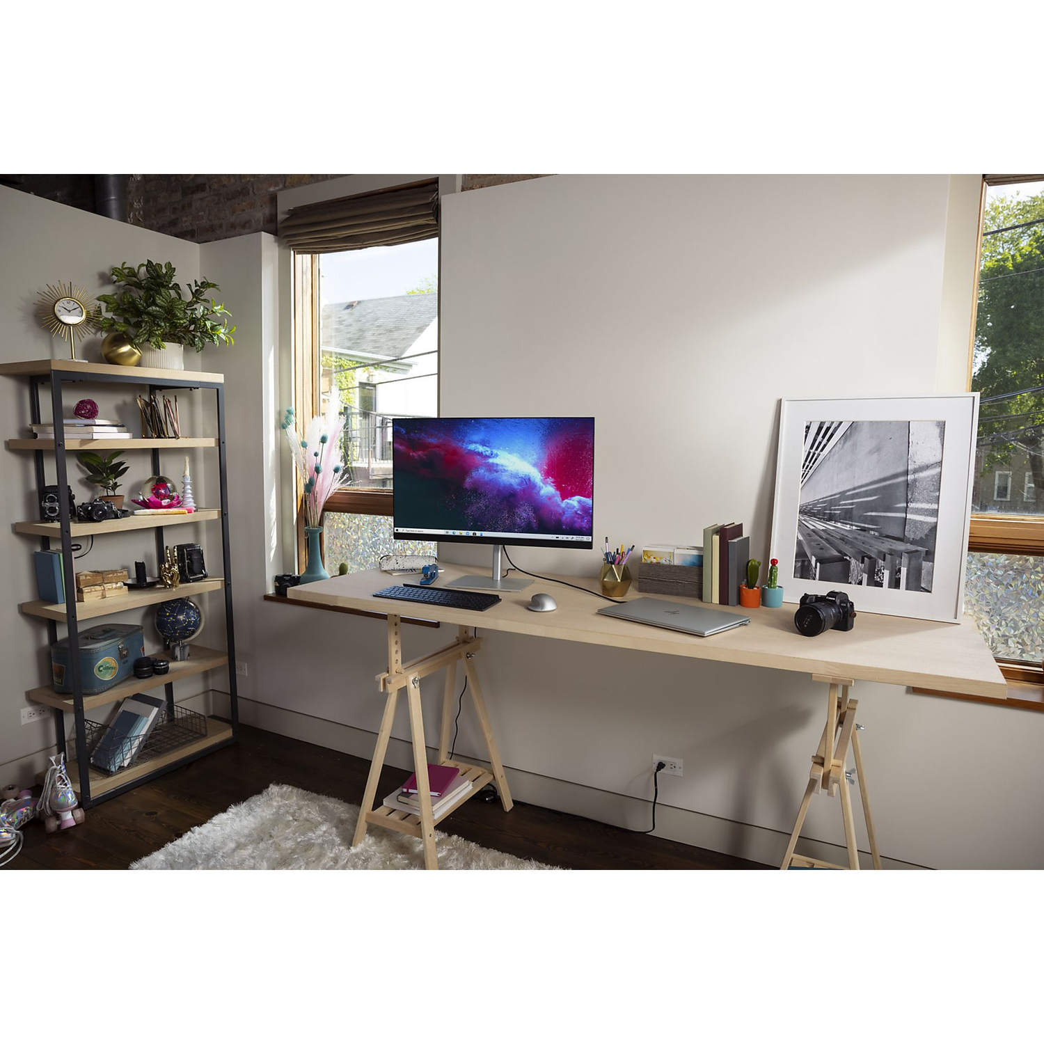 Bureaublad met computermonitor, laptop en diverse decoratieve items. Een raam op de achtergrond.