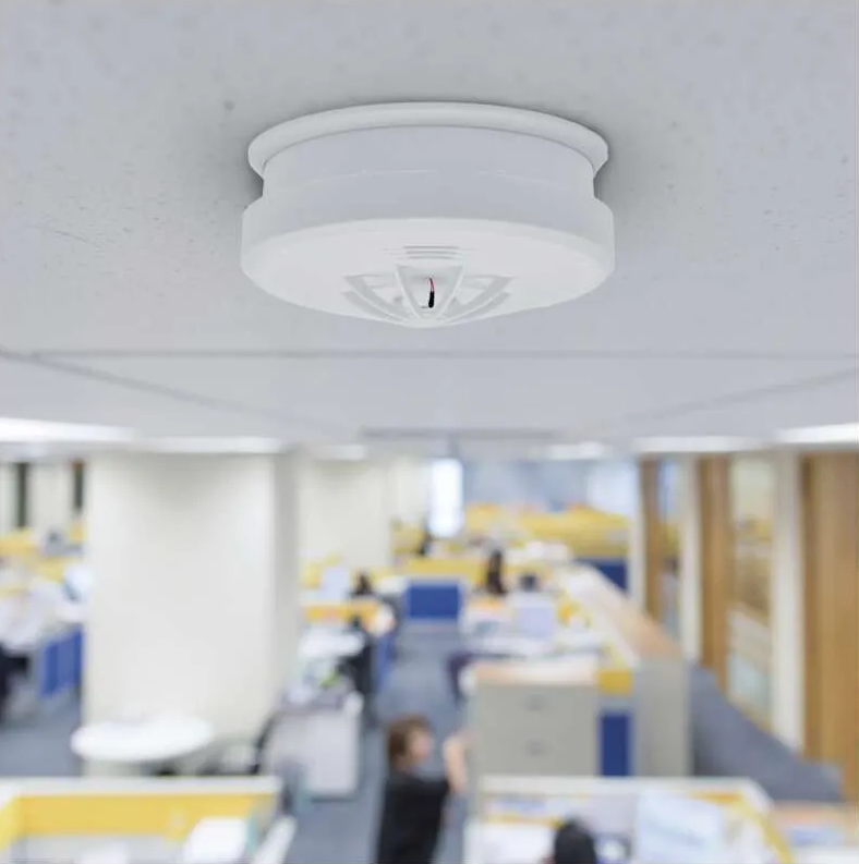 Biały czujnik dymu jest zamontowany na białym suficie w biurze. Ludzie są rozmyci w tle.