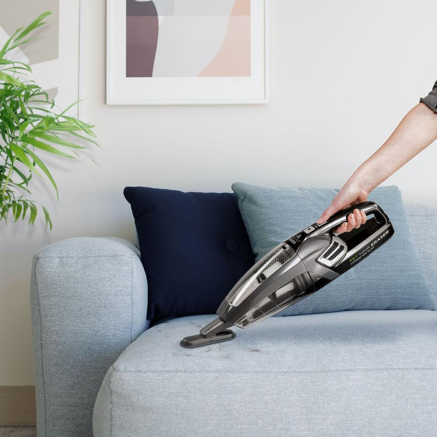 Osoba odkurzająca jasnoniebieską sofę szaro-czarnym odkurzaczem ręcznym.