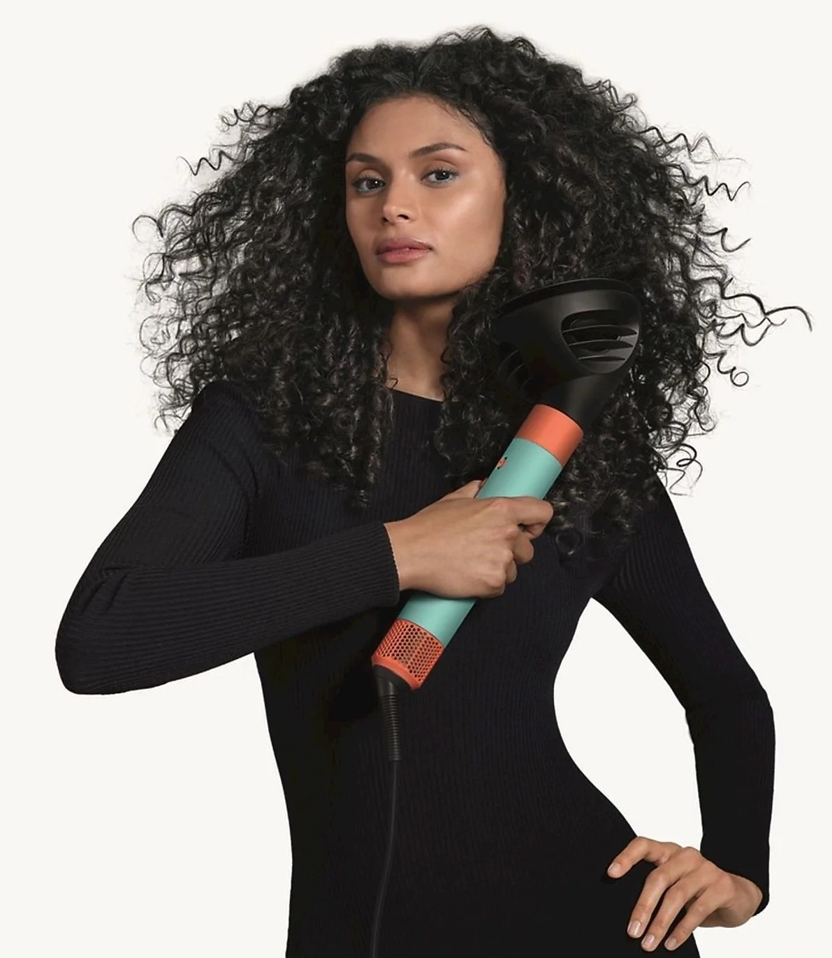 Kobieta z kręconymi włosami trzyma turkusowo-pomarańczową suszarkę do włosów, patrząc w kamerę.