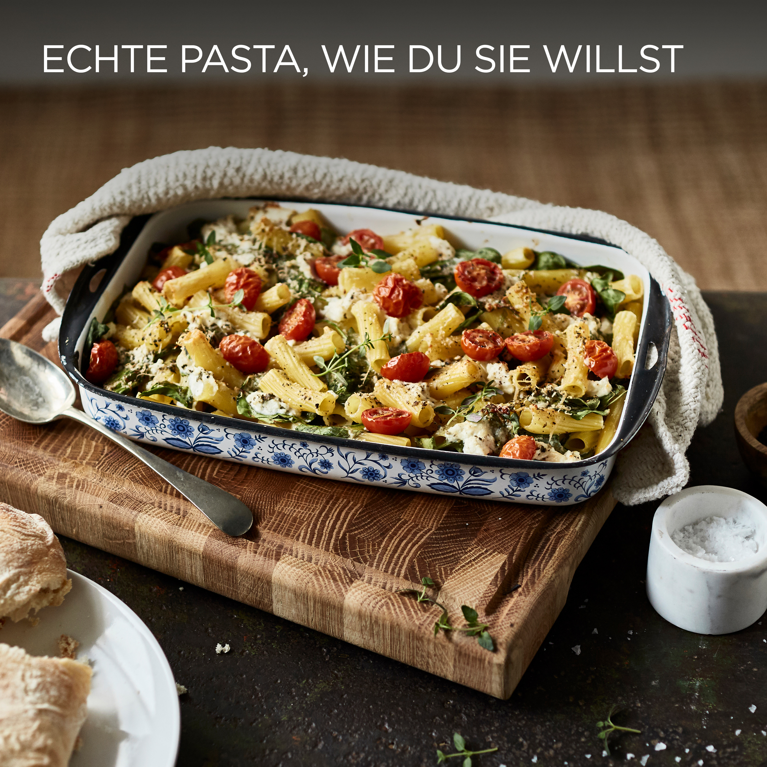 Gebackenes Pastagericht mit Tomaten, Kräutern und Käse auf einem Holzbrett, serviert mit Brot und Salz.