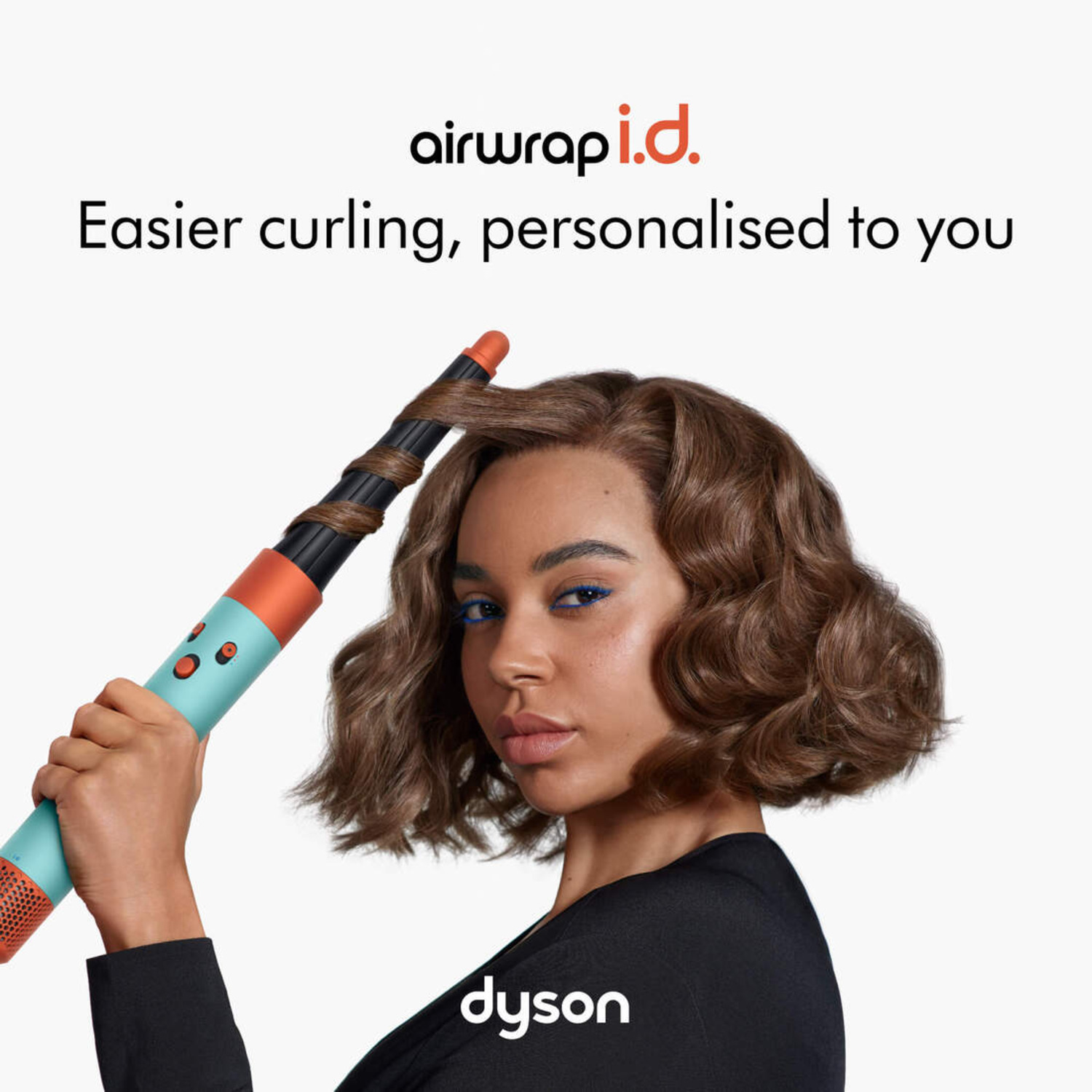 A woman with wavy brown hair is styling her hair with a curling iron, white background.