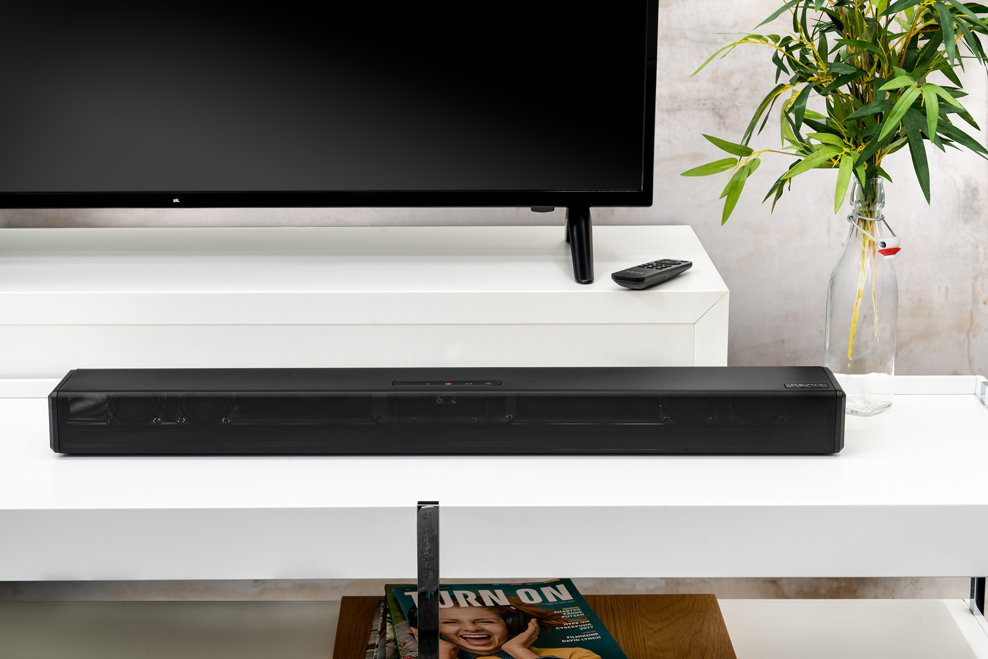 Fekete soundbar fehér polcon, fekete TV alatt. Zöld növényekkel teli váza jobbra.