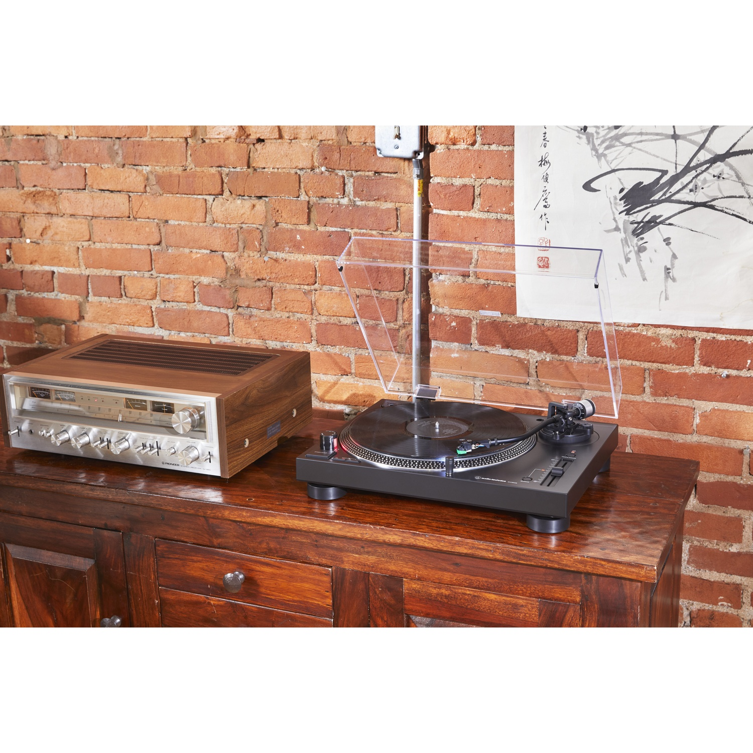 Un giradischi e un ricevitore su un mobile in legno contro un muro di mattoni e un'opera d'arte.