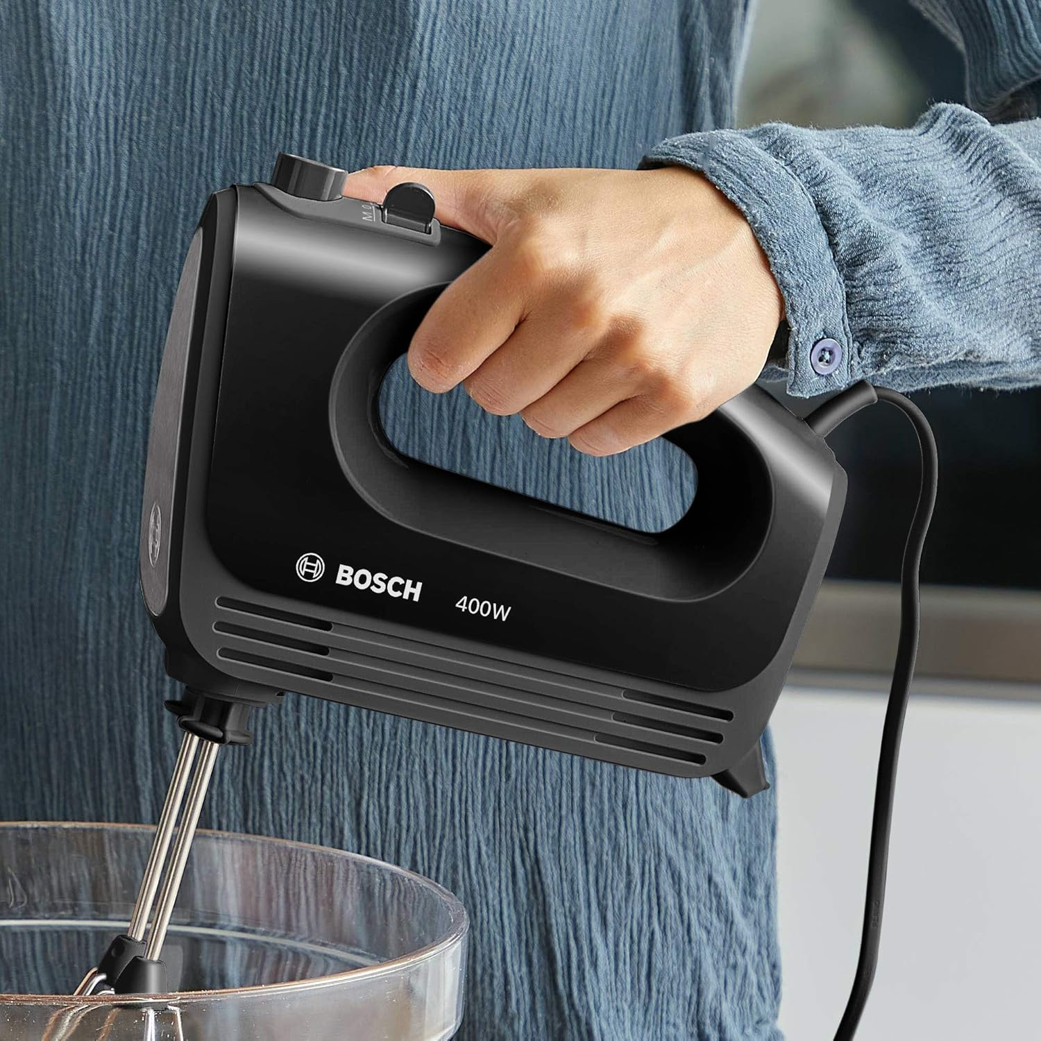 Ręka osoby trzymająca czarny mikser ręczny Bosch nad szklaną miską.